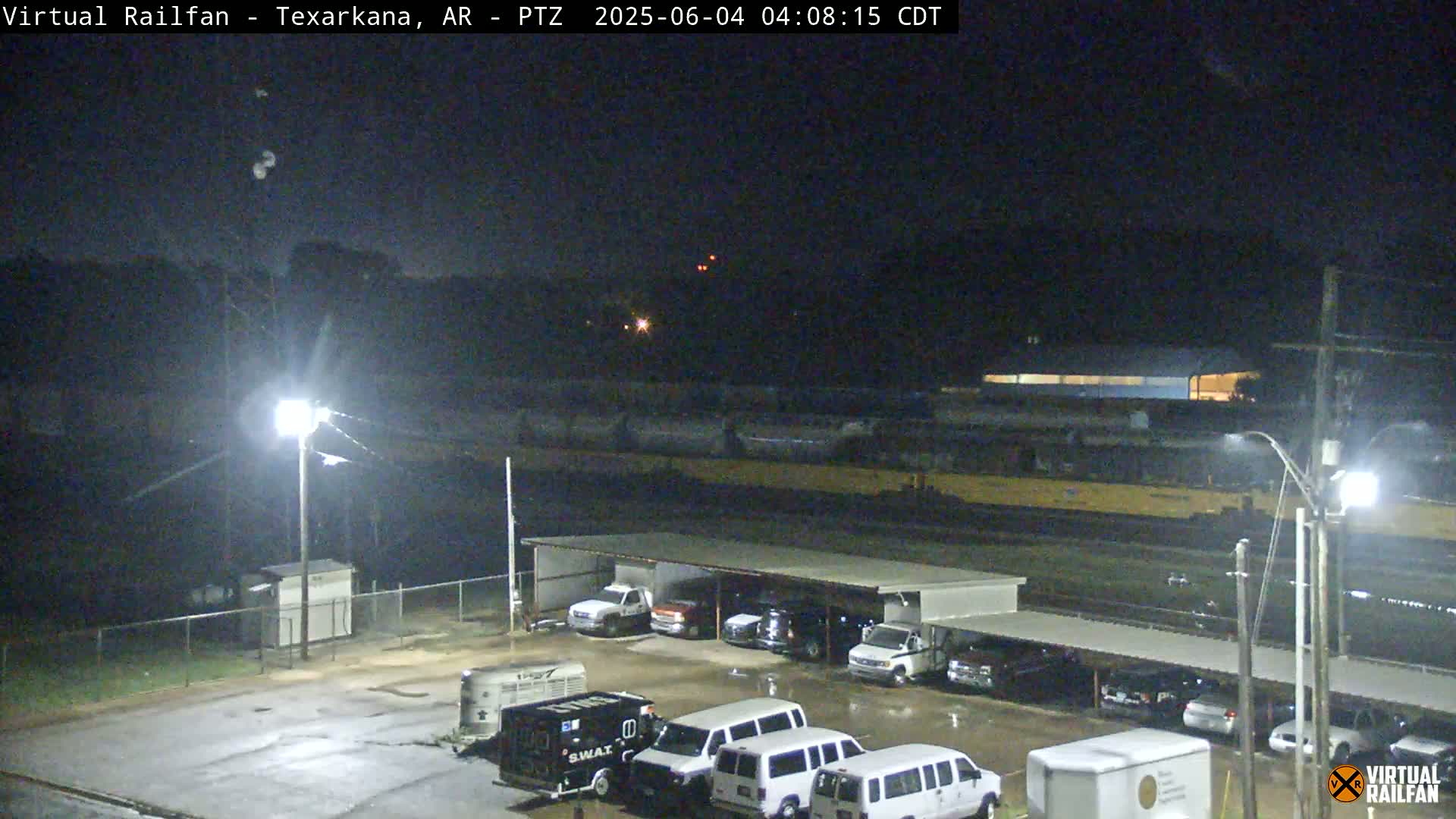 A nighttime view of a railway yard with several vehicles parked under a shelter, illuminated by artificial lights under a clear, dark sky.