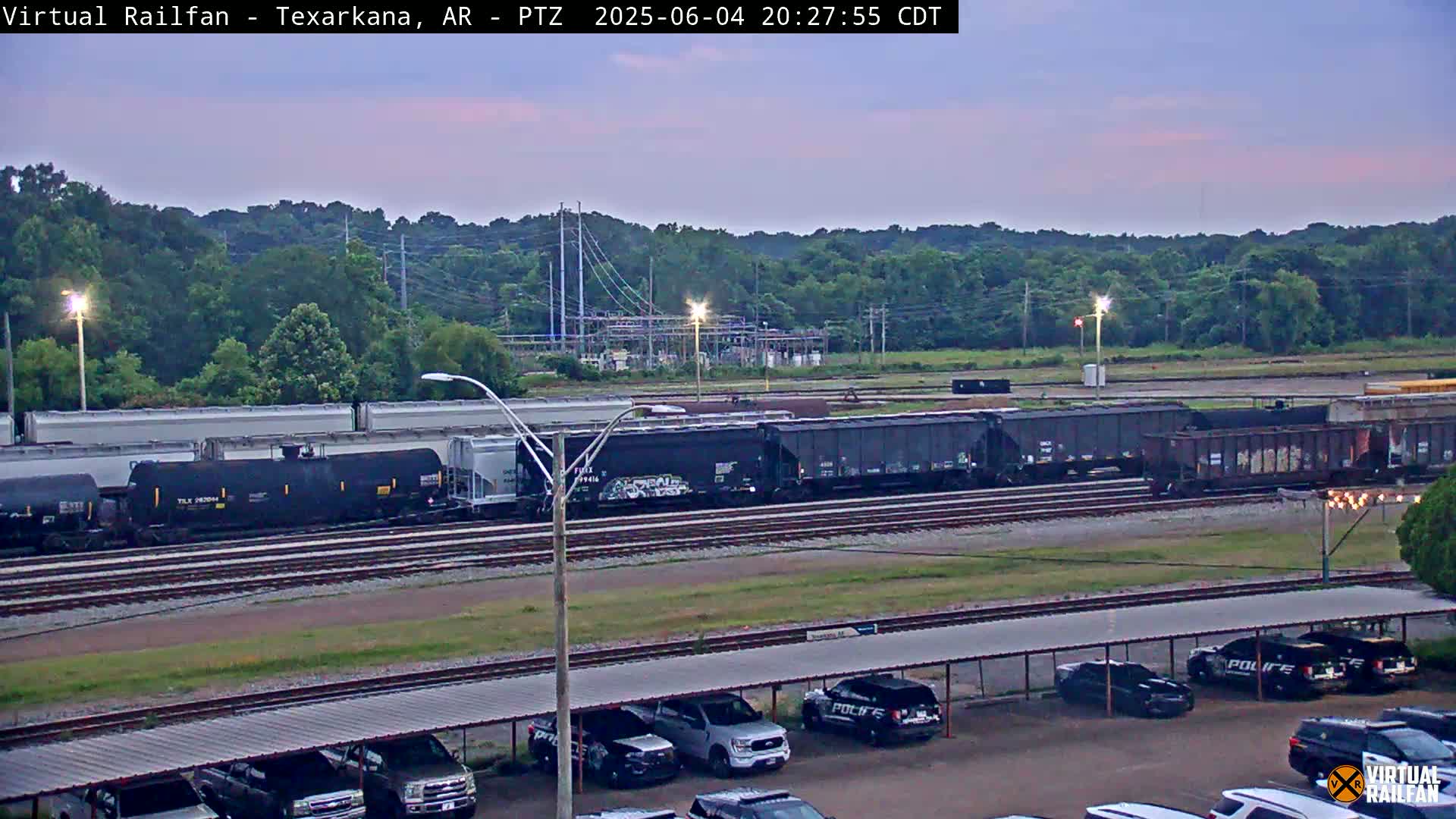 A long freight train sits on railroad tracks in a parking lot under a twilight sky, with several police cars parked nearby.