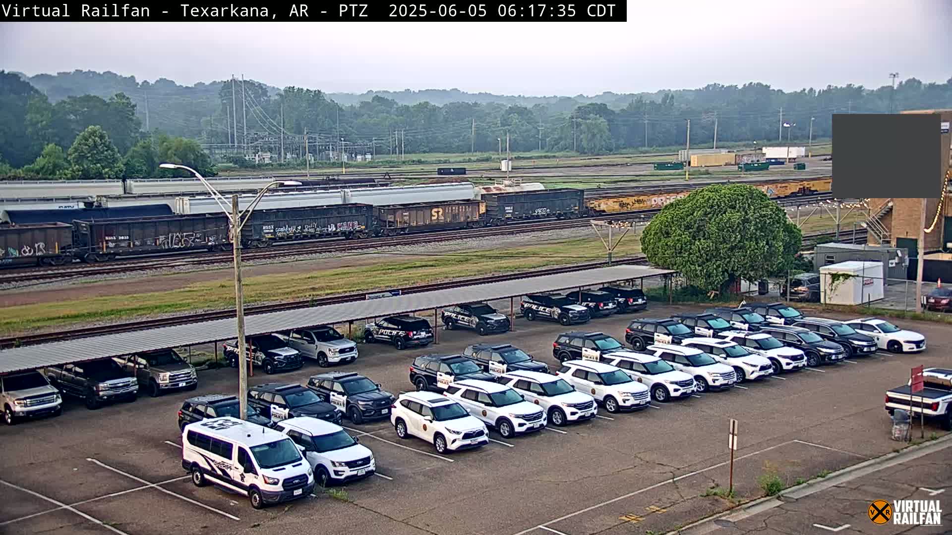 A row of police vehicles is parked under a covered structure near a railway with a freight train in the background, on a hazy day.