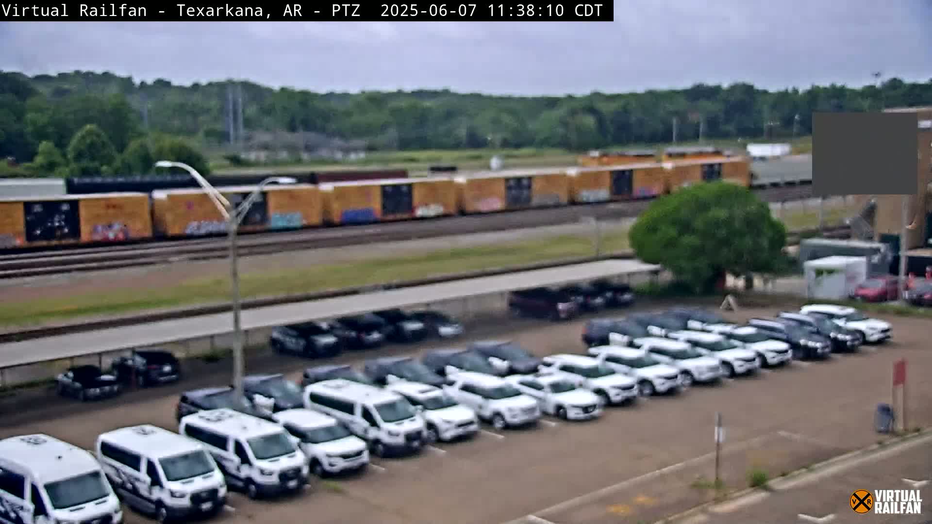 A train of boxcars sits on tracks behind a parking lot filled with mostly white vans and SUVs under an overcast sky.