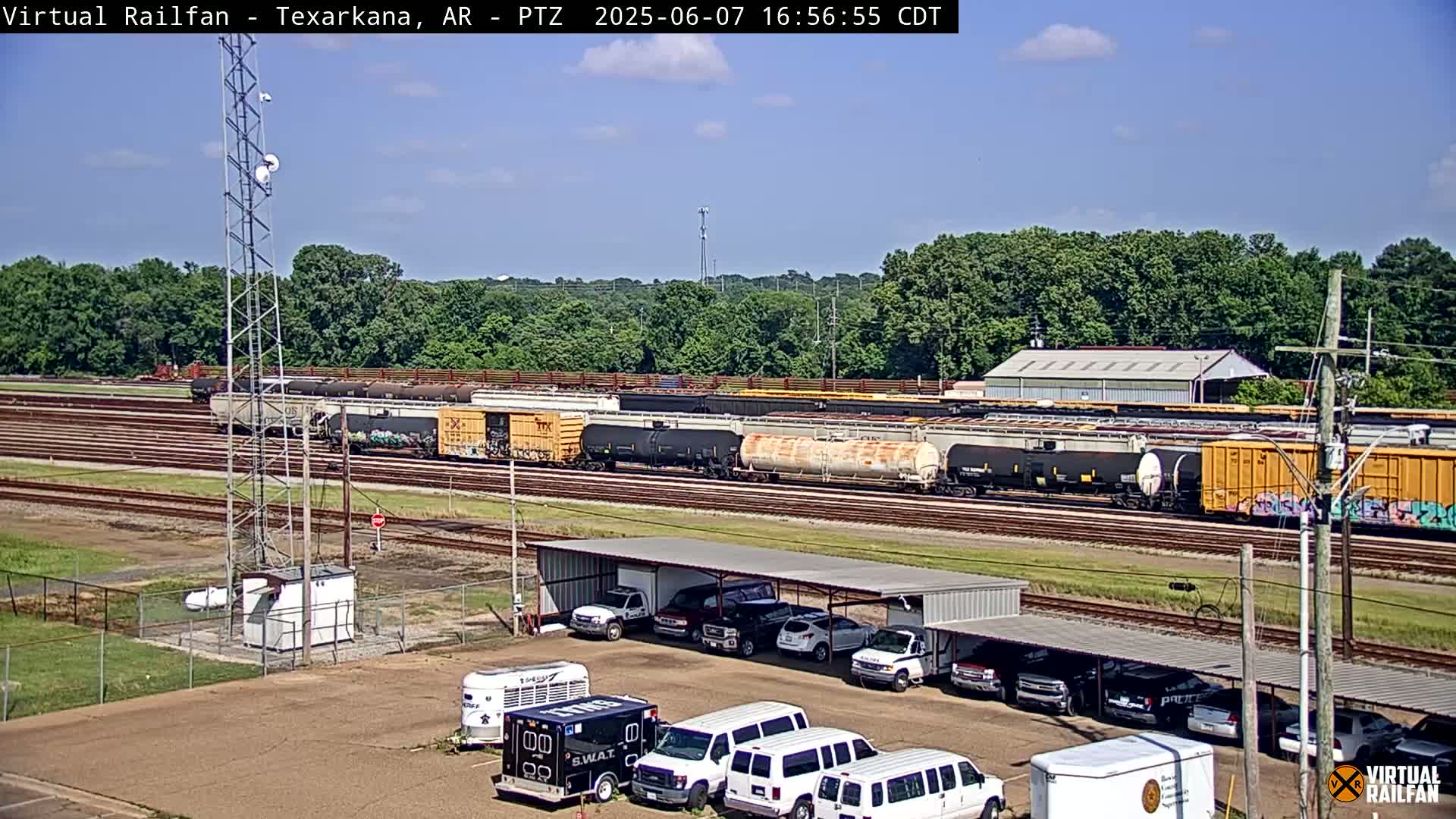 A sunny day reveals a train yard with several freight cars parked on multiple tracks, a parking lot with numerous vehicles under a covered structure, and a tall communication tower.
