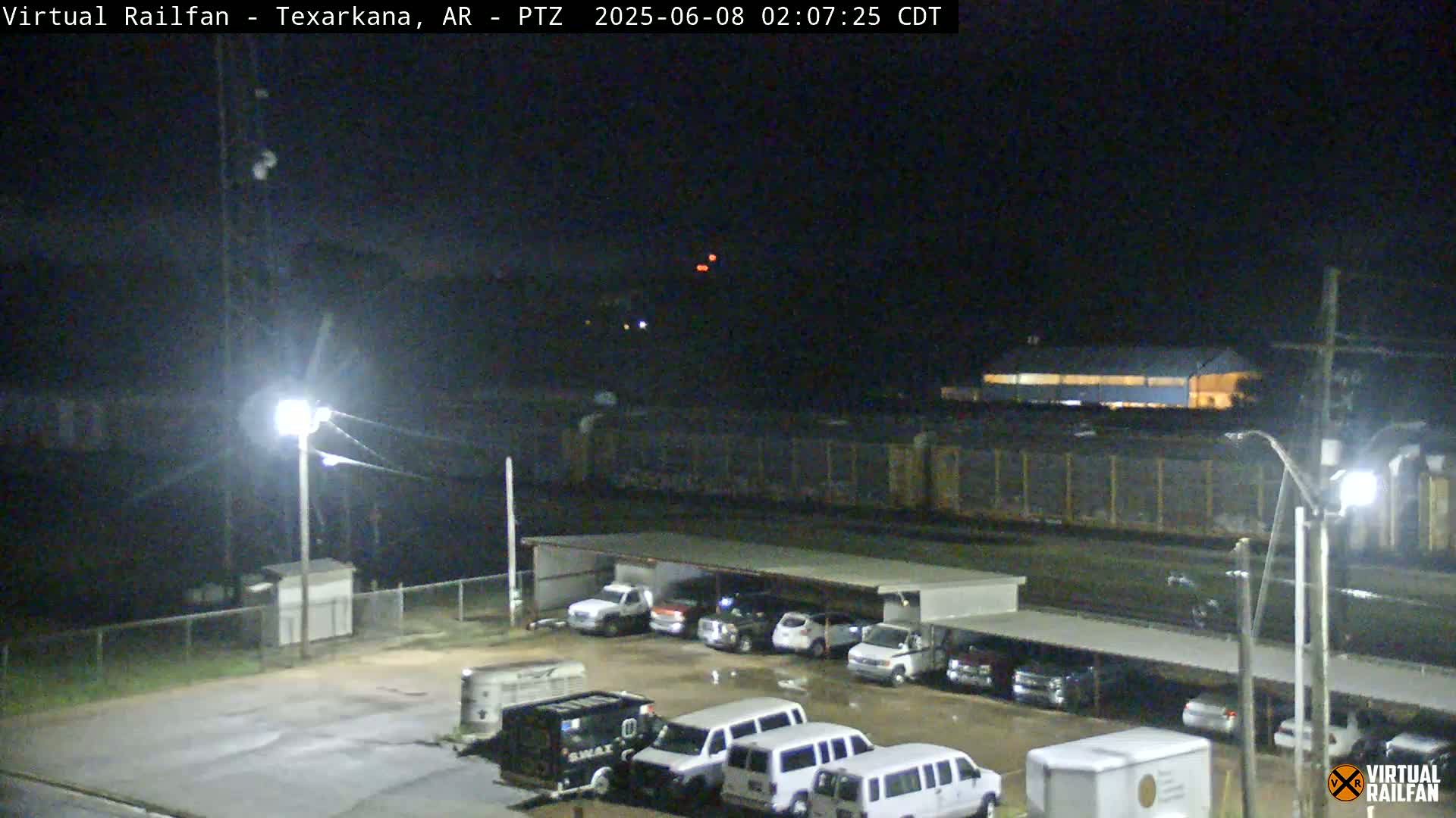 A nighttime view of a parking lot with several vehicles under a covered structure, next to a long train stopped on tracks.