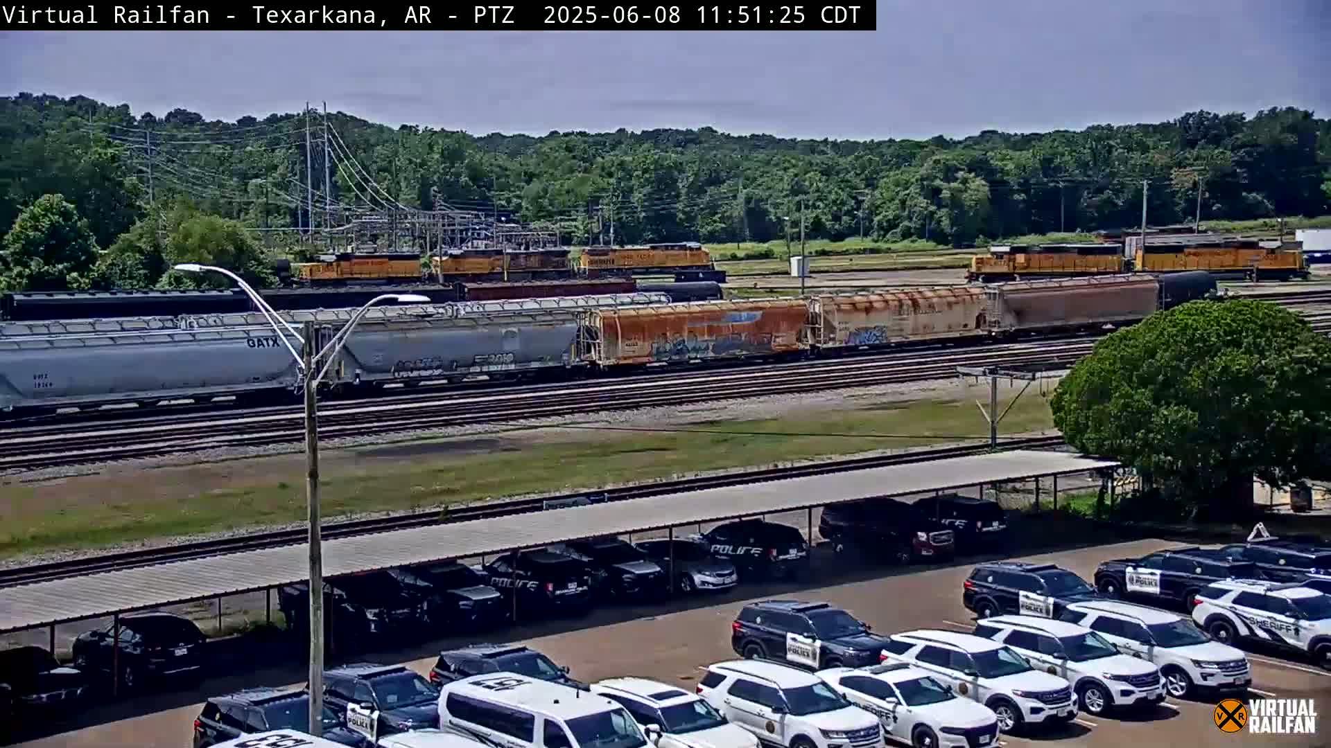 A freight train passes by a parking lot full of police vehicles under a covered structure on a sunny day.