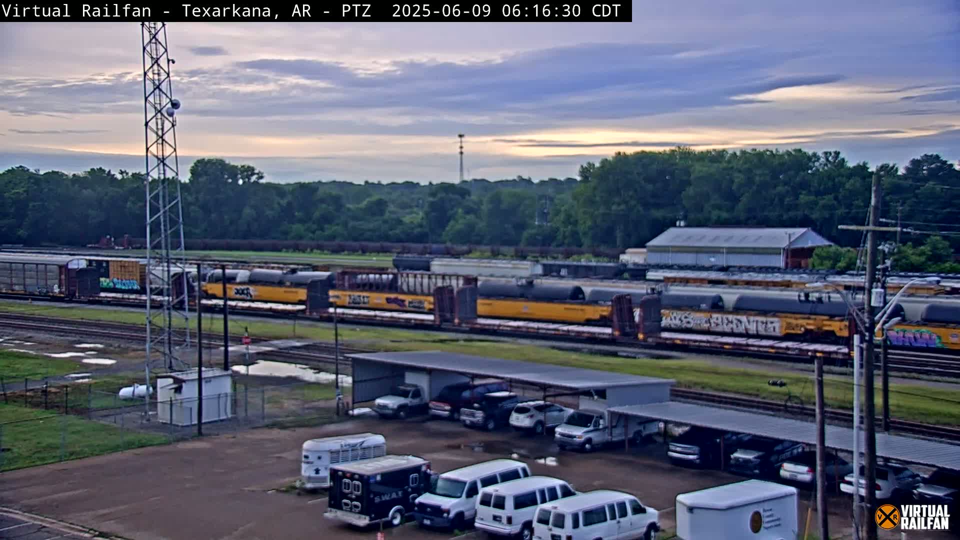A long freight train passes by a parking lot with several vans and other vehicles under a covered shelter, under a partly cloudy sunrise sky.