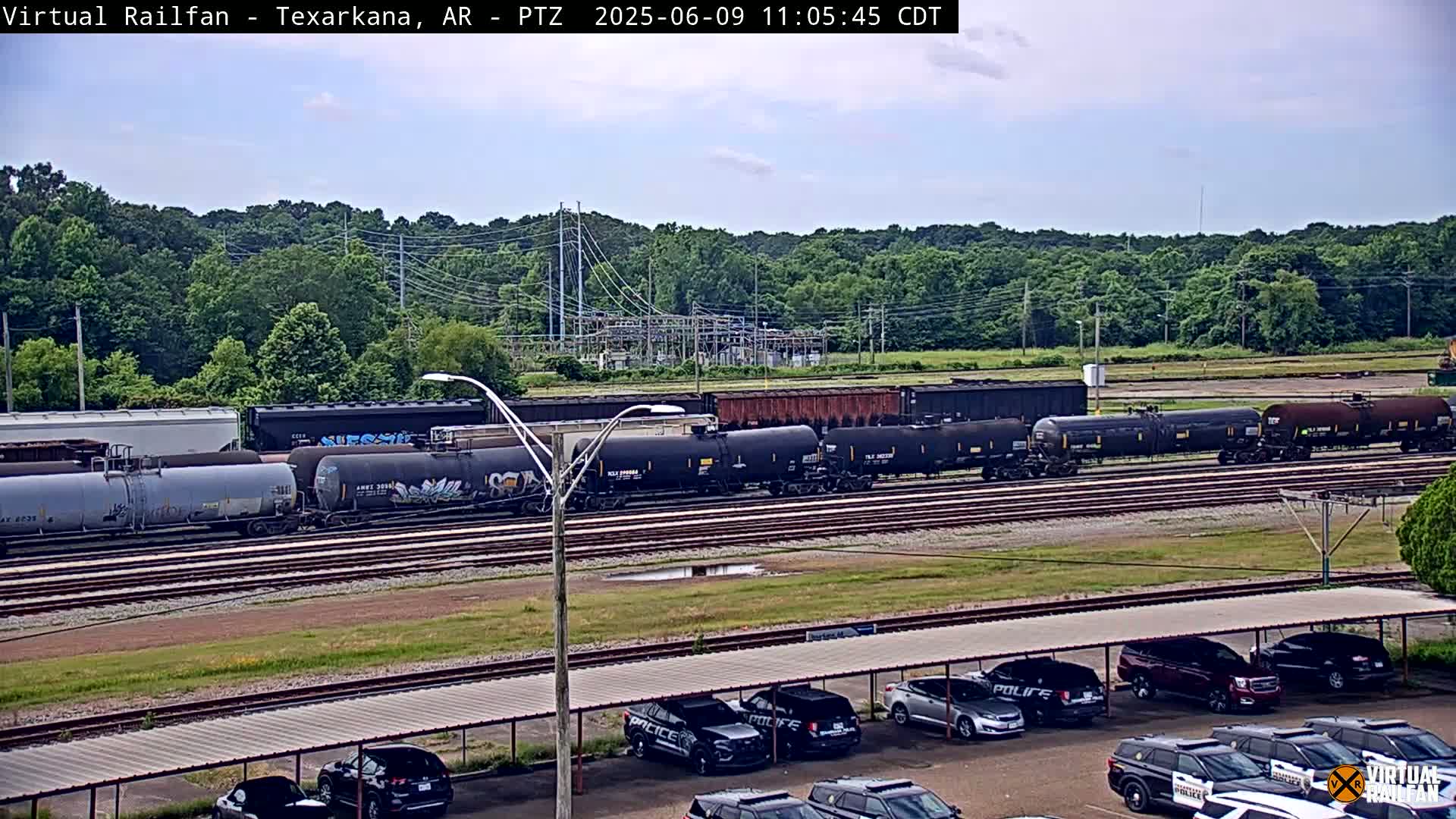 A long freight train of tanker cars sits on a railway track in front of a wooded area under a partly cloudy sky.
