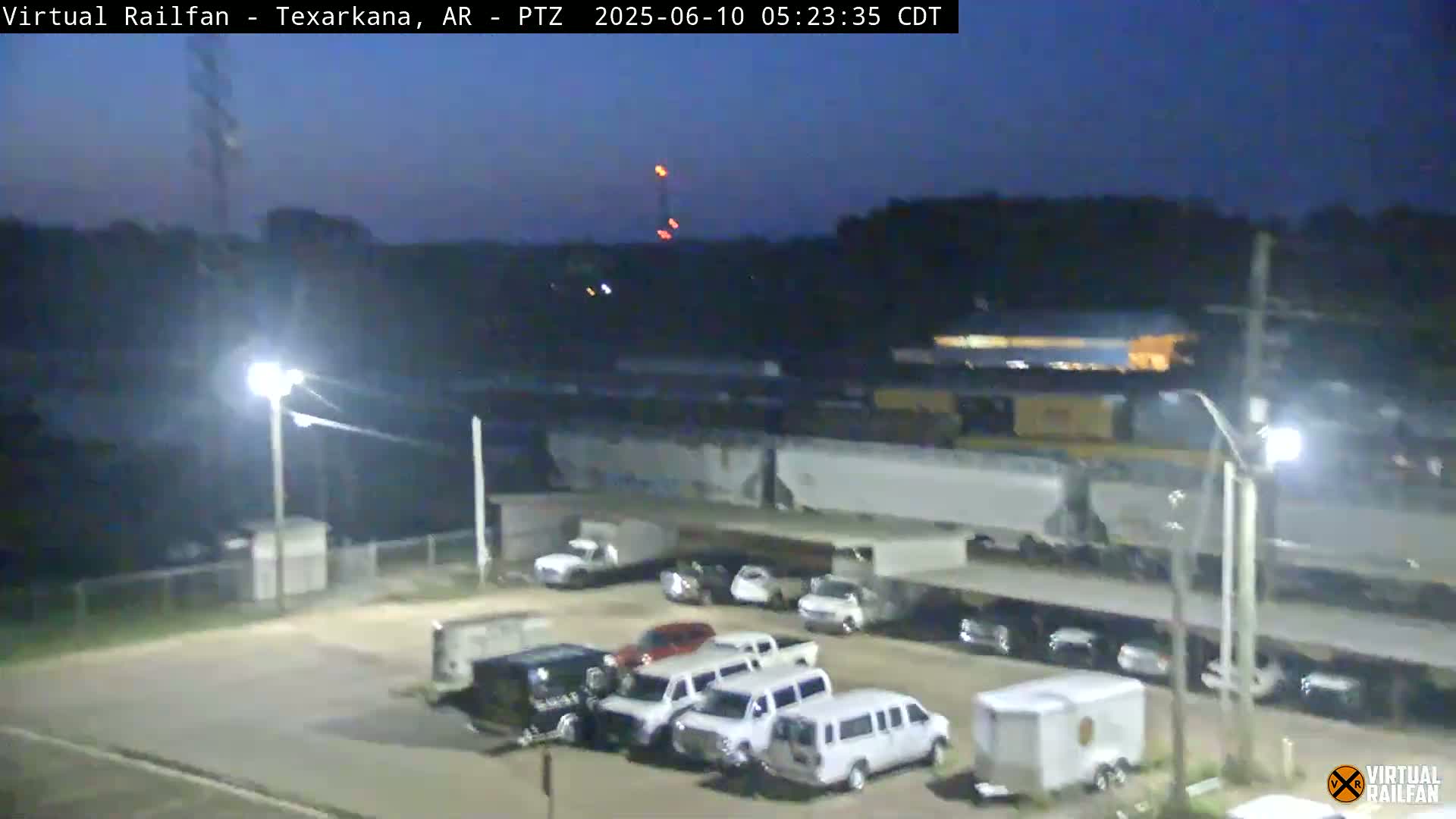 A nighttime, dimly lit view of a parking lot with several vehicles, next to a railway with a train passing by under a covered structure.