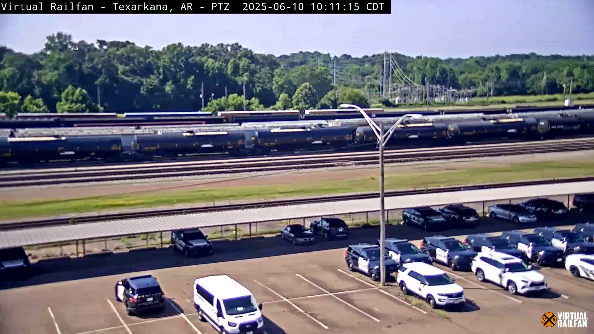 A long freight train passes by a parking lot filled with police vehicles under a sunny sky.