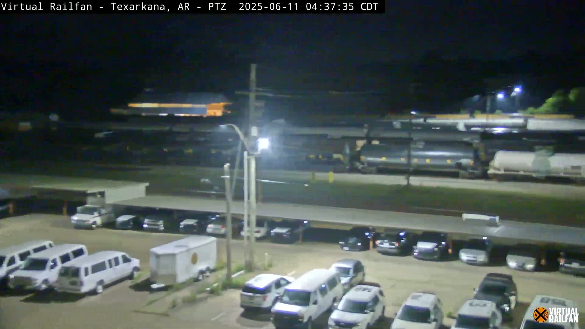 A nighttime view of a parking lot filled with cars and vans, adjacent to a train carrying tanker cars.