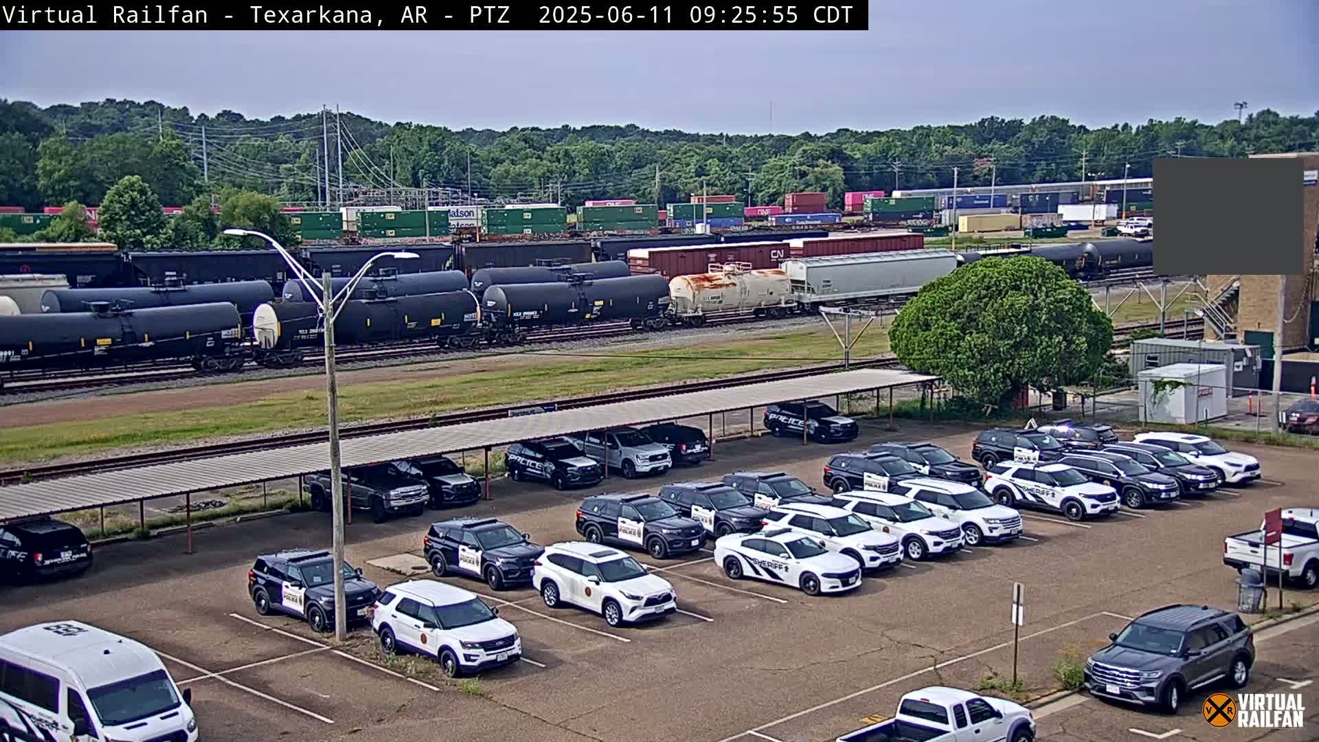 Under a mostly clear sky, a train carrying tanker cars is parked near a lot full of police vehicles.