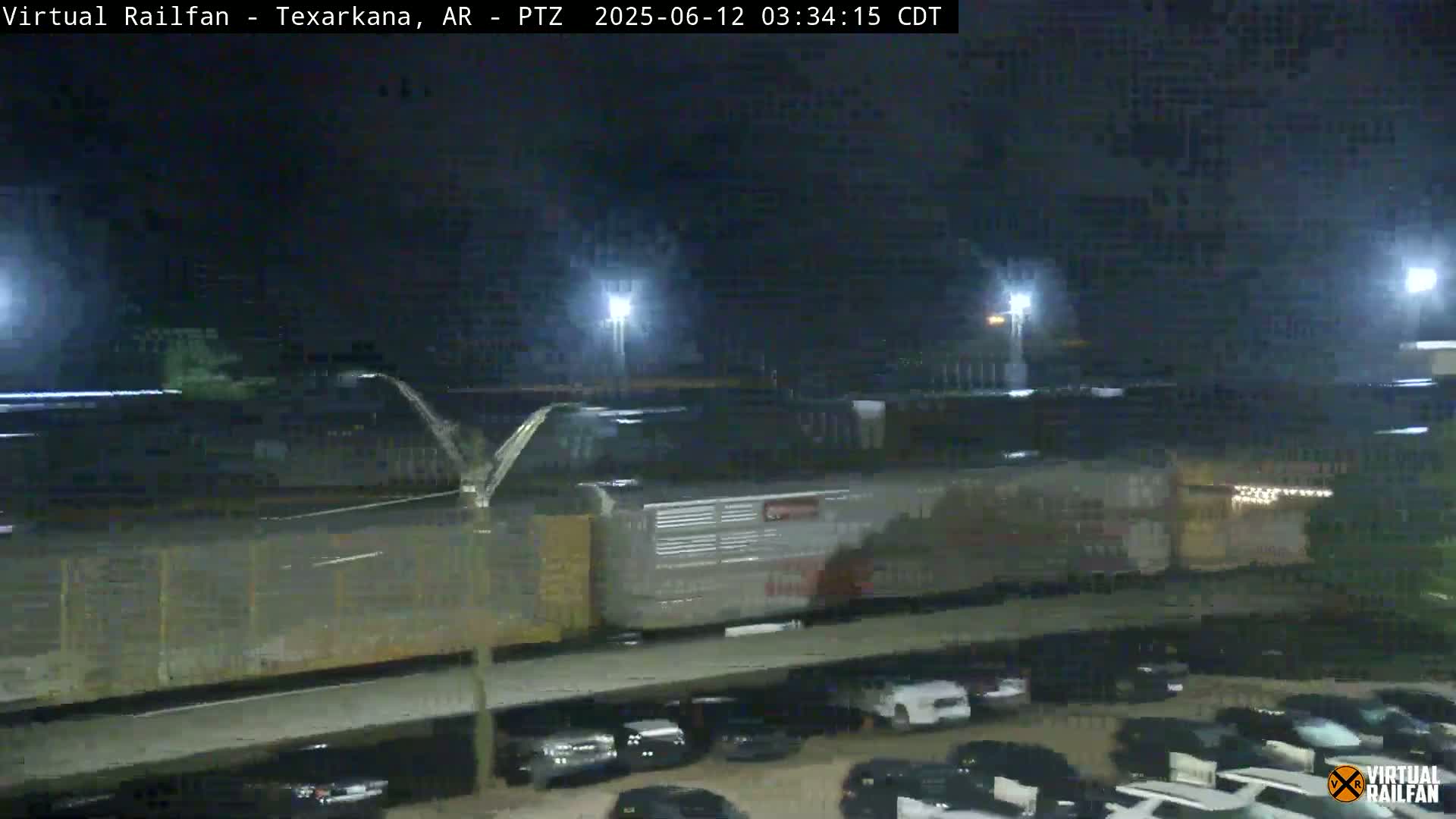 A long freight train is moving through a rail yard at night, with a parking lot visible in the foreground.