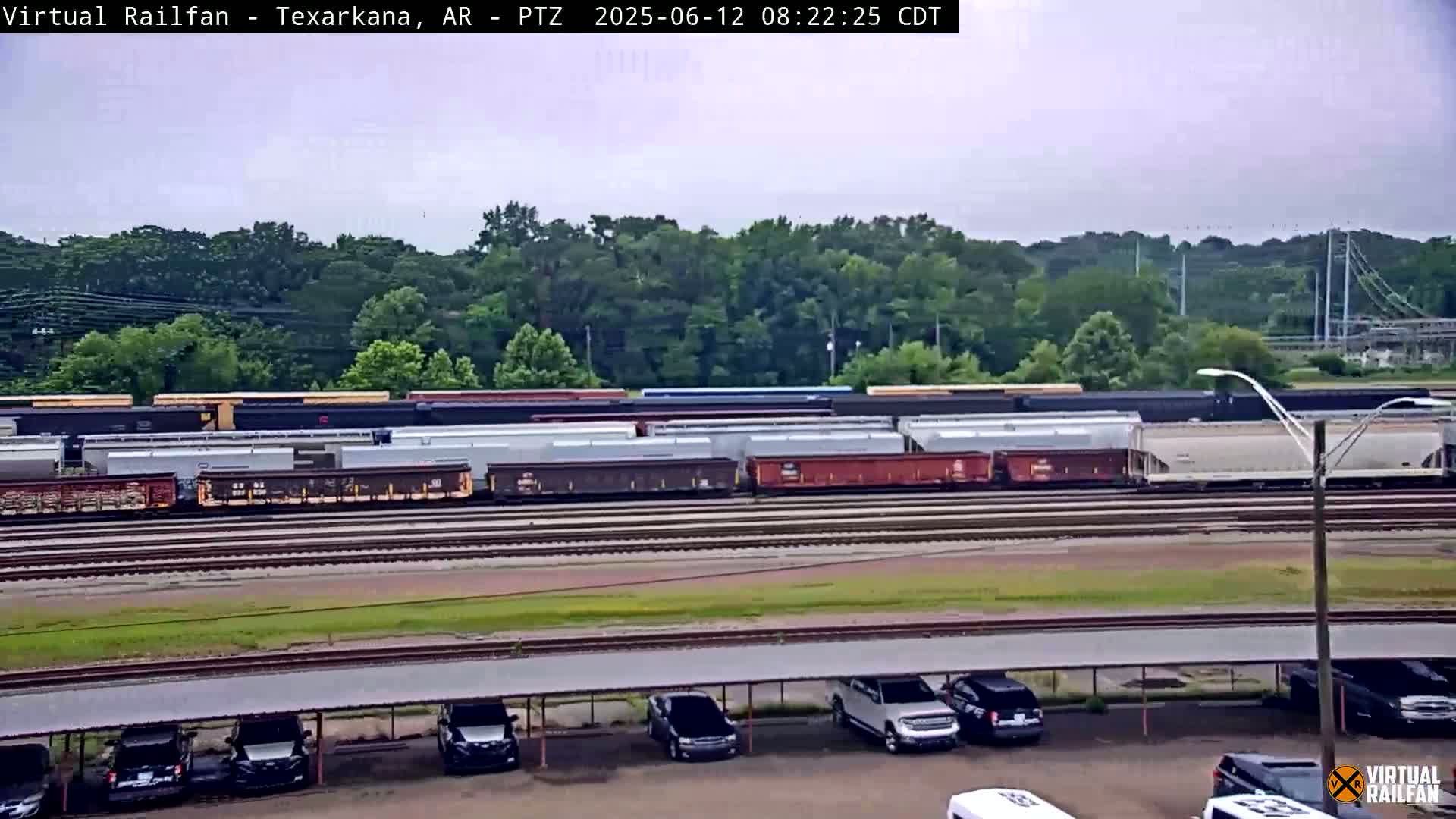 A long train of freight cars sits on a railway track next to a parking lot under an overcast sky.