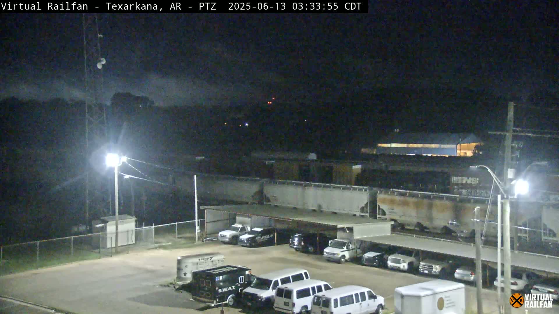 A nighttime view of a rail yard with several parked vehicles under a covered area, a train in the background, and a partly cloudy sky.