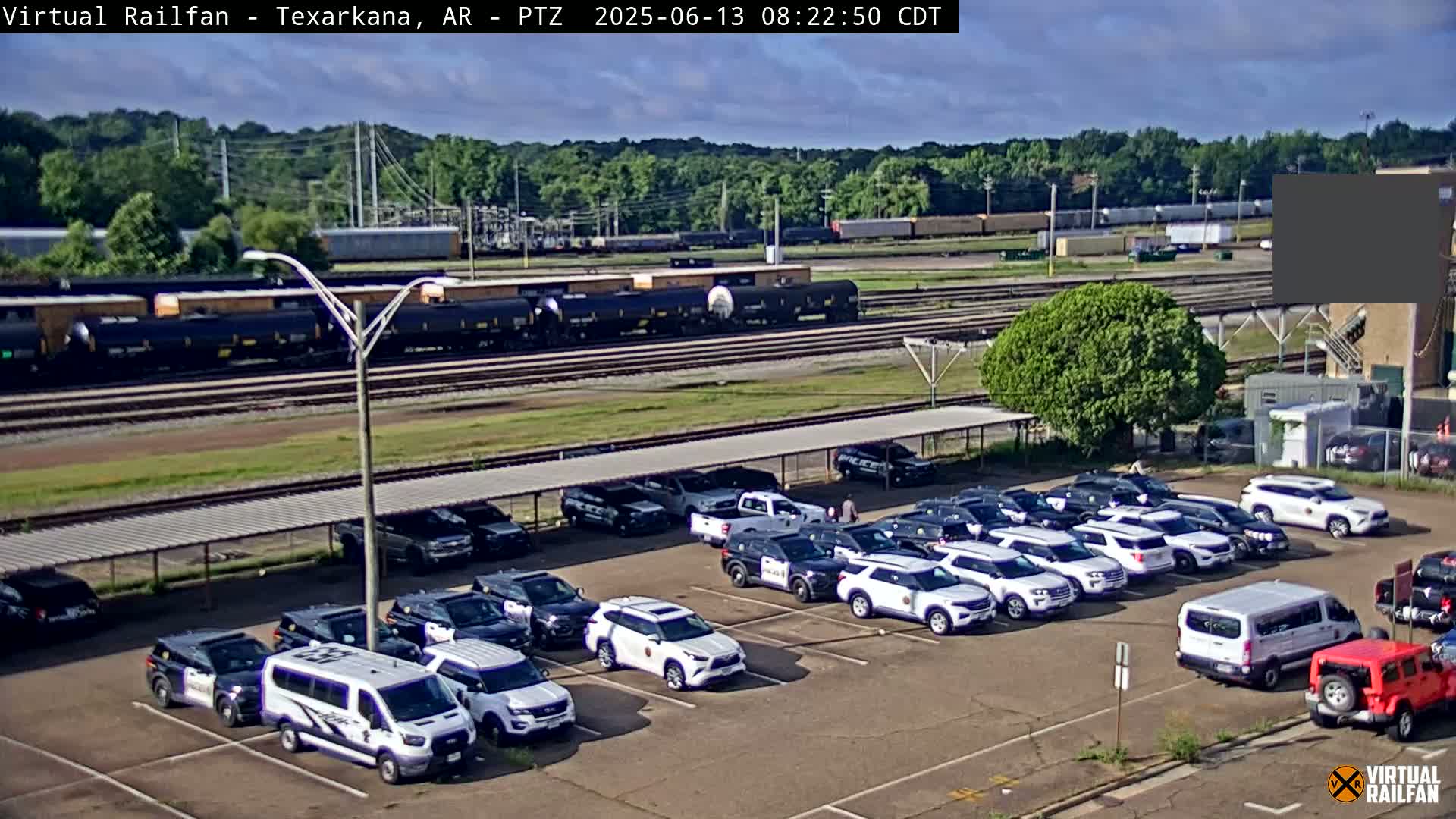 A parking lot filled with numerous white police vehicles is situated near a railway track where a freight train is passing under partly cloudy skies.