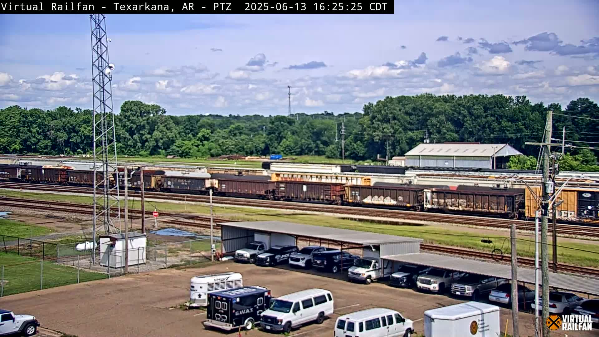 A long freight train sits on the tracks under a partly cloudy sky, next to a parking lot with several vehicles under a covered structure.