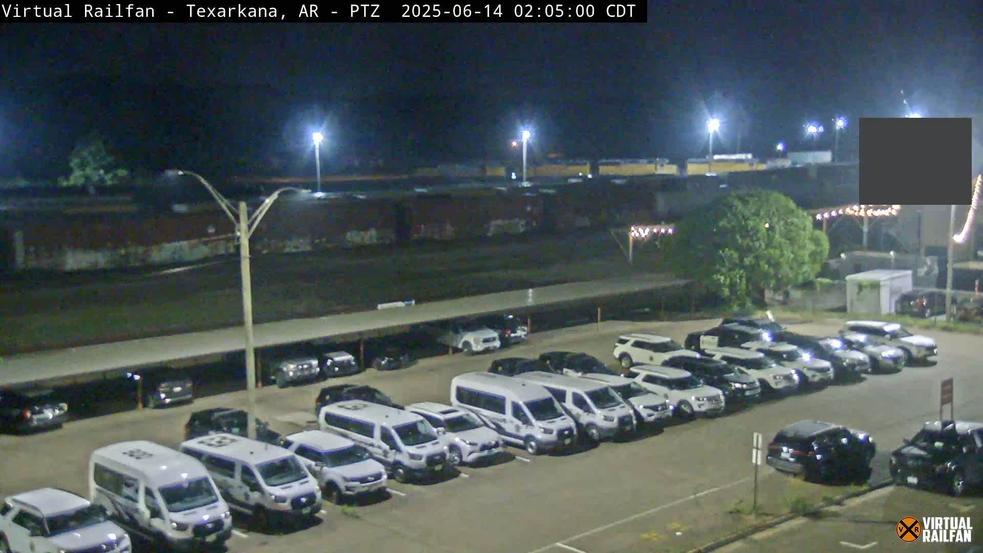 A nighttime view of a parking lot filled with white vans and SUVs, next to a train car under a covered area.