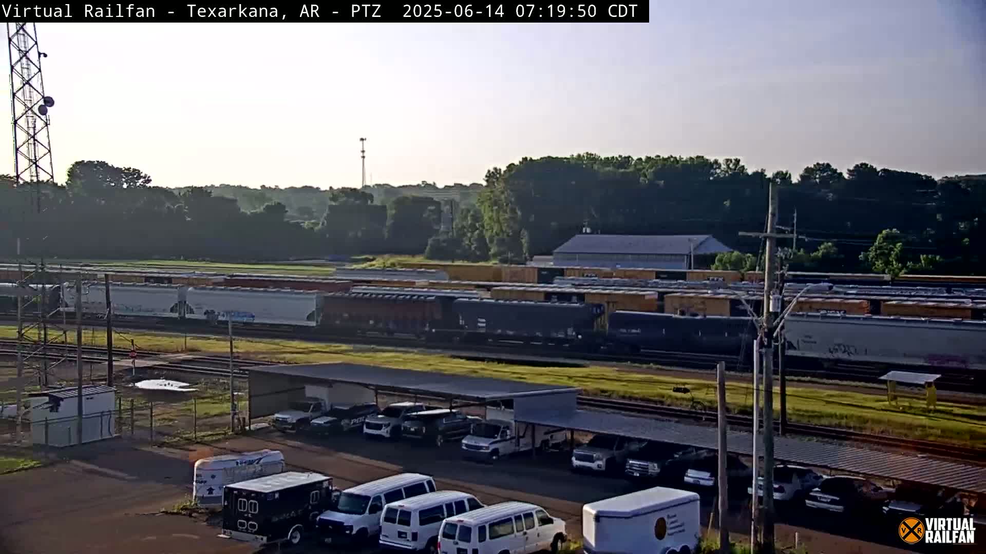 A long freight train passes by a parking lot filled with various vehicles under a covered structure, on a partly sunny day.