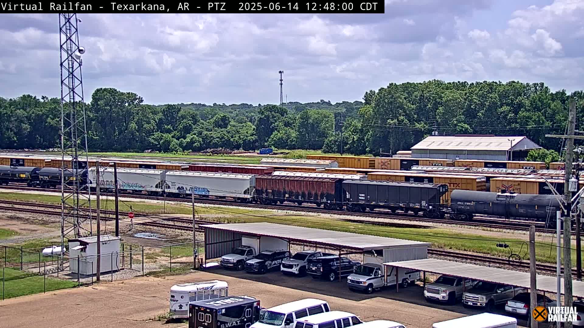 A long freight train sits on railway tracks under a partly cloudy sky, next to a parking lot with several vehicles and a building.