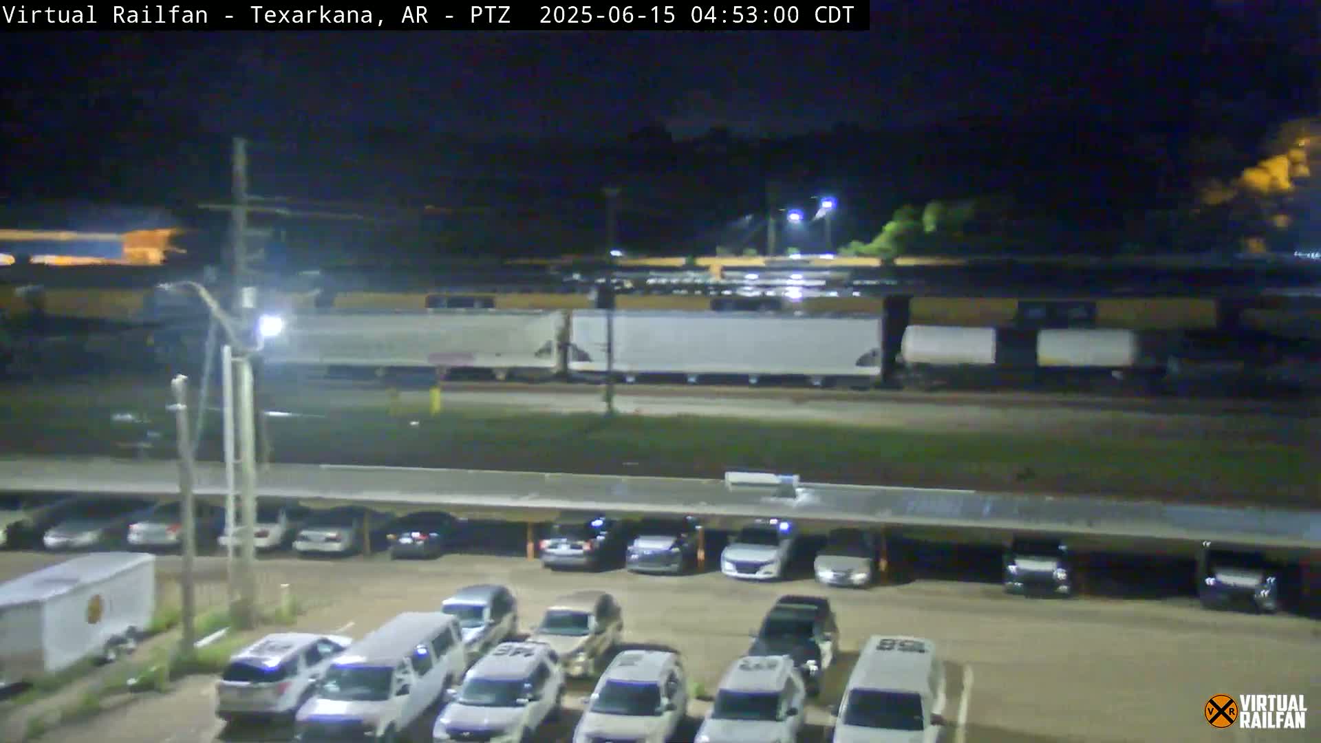 At night, a long freight train passes by a parking lot filled with cars under a covered structure.