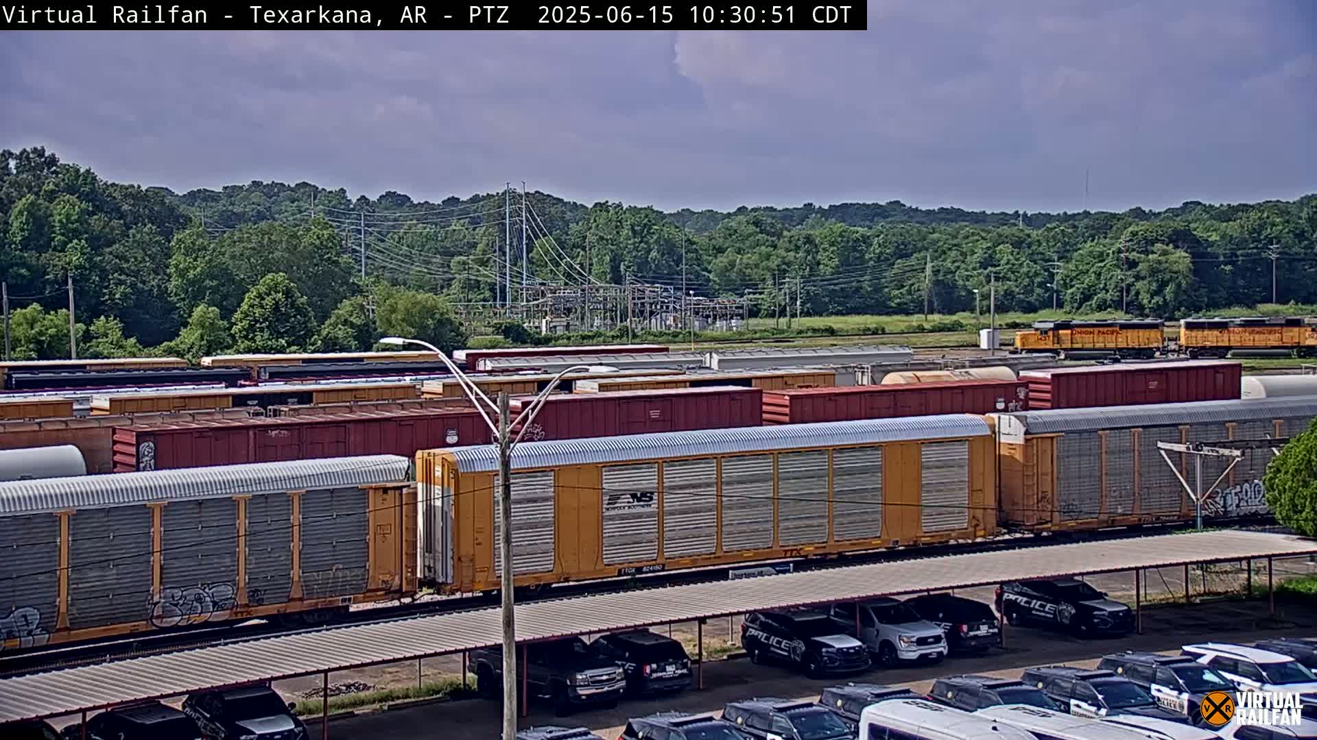 Numerous freight cars are parked in a rail yard under a partly cloudy sky, with a line of police vehicles visible in a parking lot in the foreground.