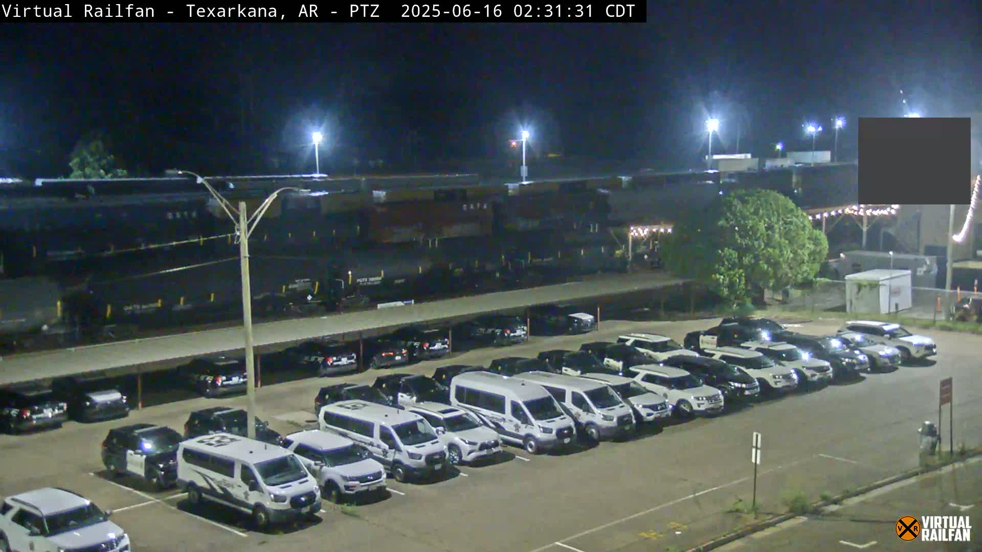 At night, a long freight train sits beside a parking lot full of mostly white vans and SUVs.