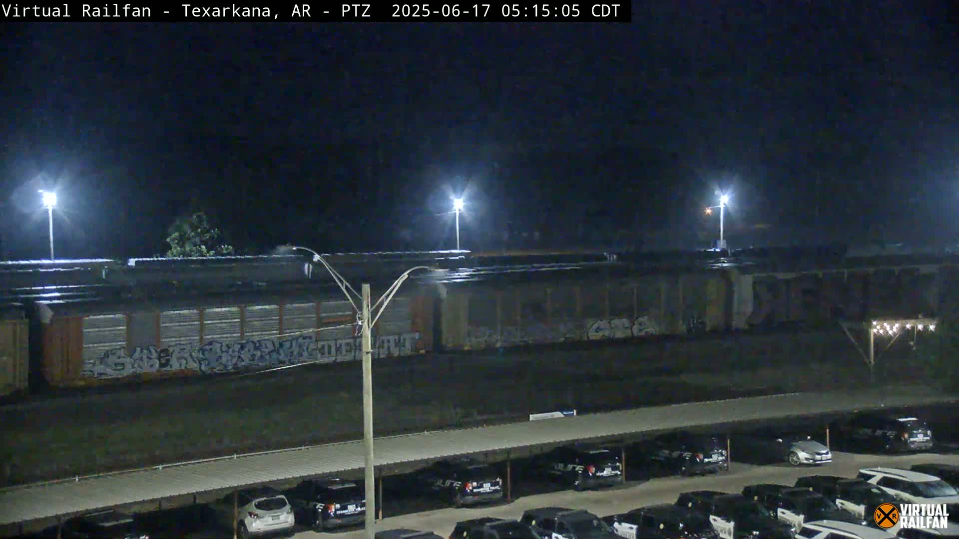 A nighttime view of a graffiti-covered train car parked next to a parking lot full of cars under a covered structure.