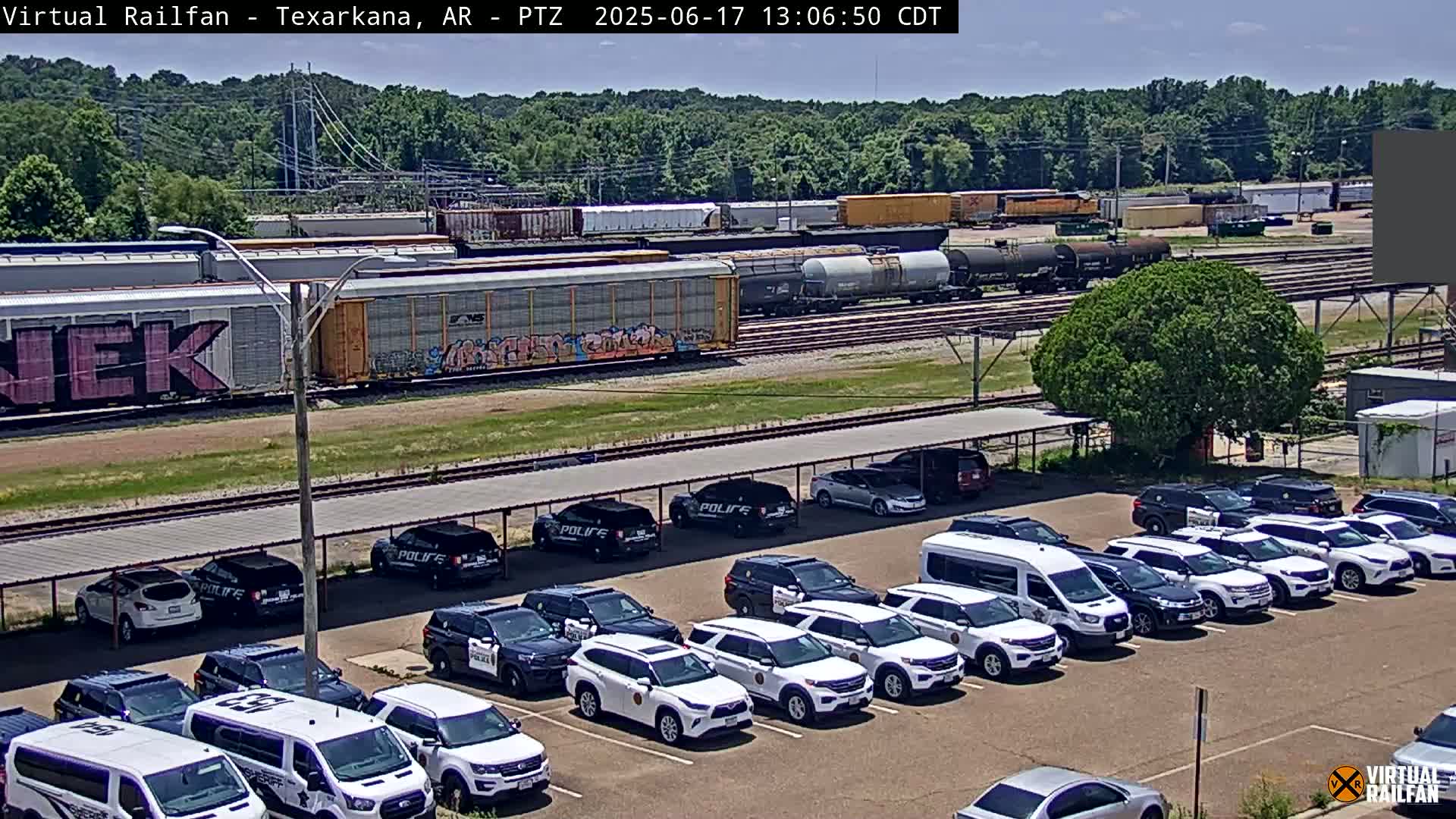 A freight train passes by a parking lot filled with police vehicles under a sunny sky.