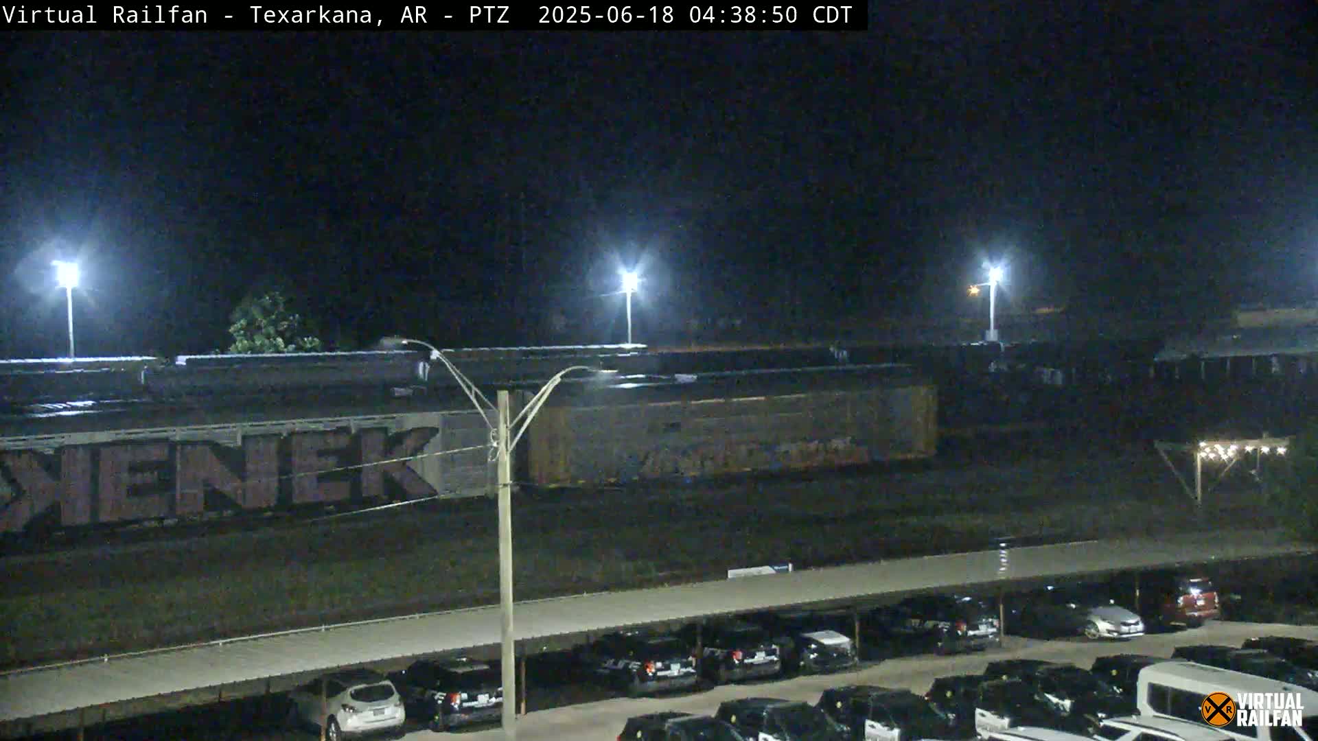 A nighttime view of a graffiti-covered train car next to a parking lot filled with cars under a covered structure.