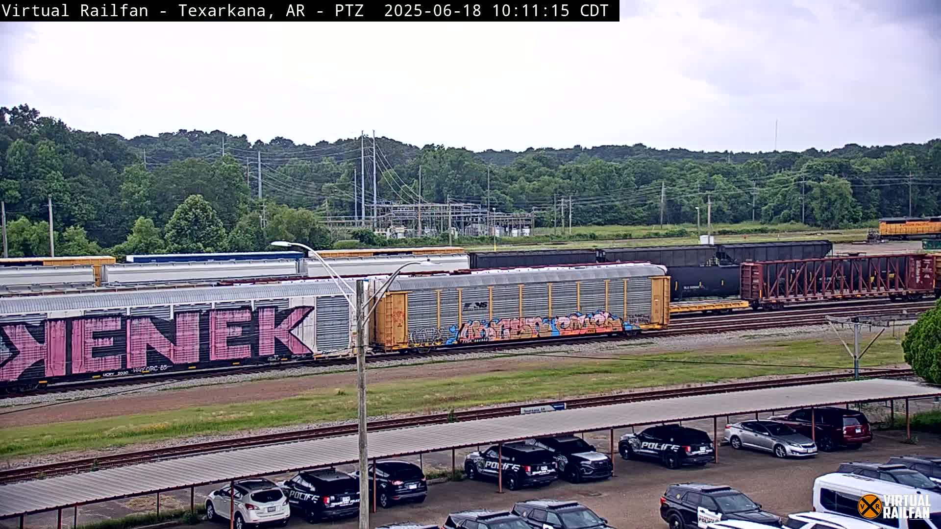 A freight train with graffiti-covered cars sits on railway tracks next to a parking lot filled with police vehicles under an overcast sky.