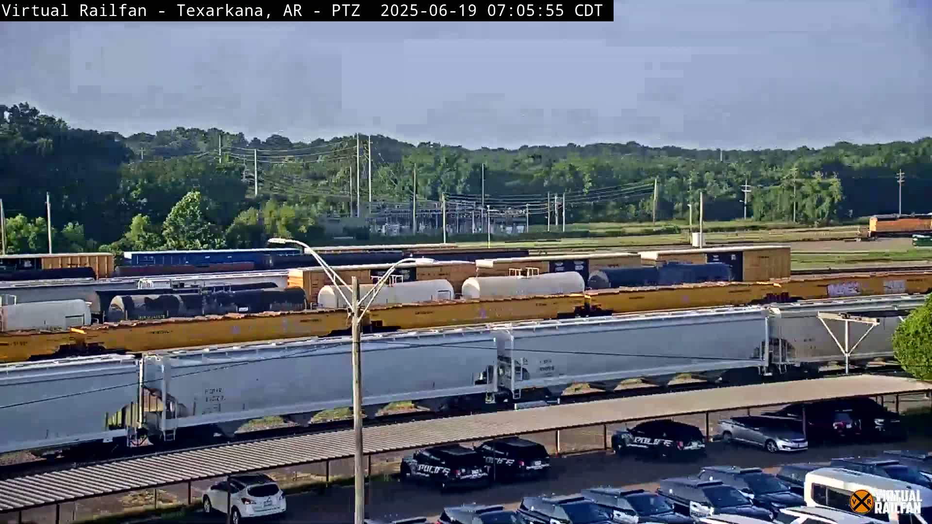 A long freight train with yellow and gray cars sits on tracks next to a parking lot full of cars under a partly cloudy sky.