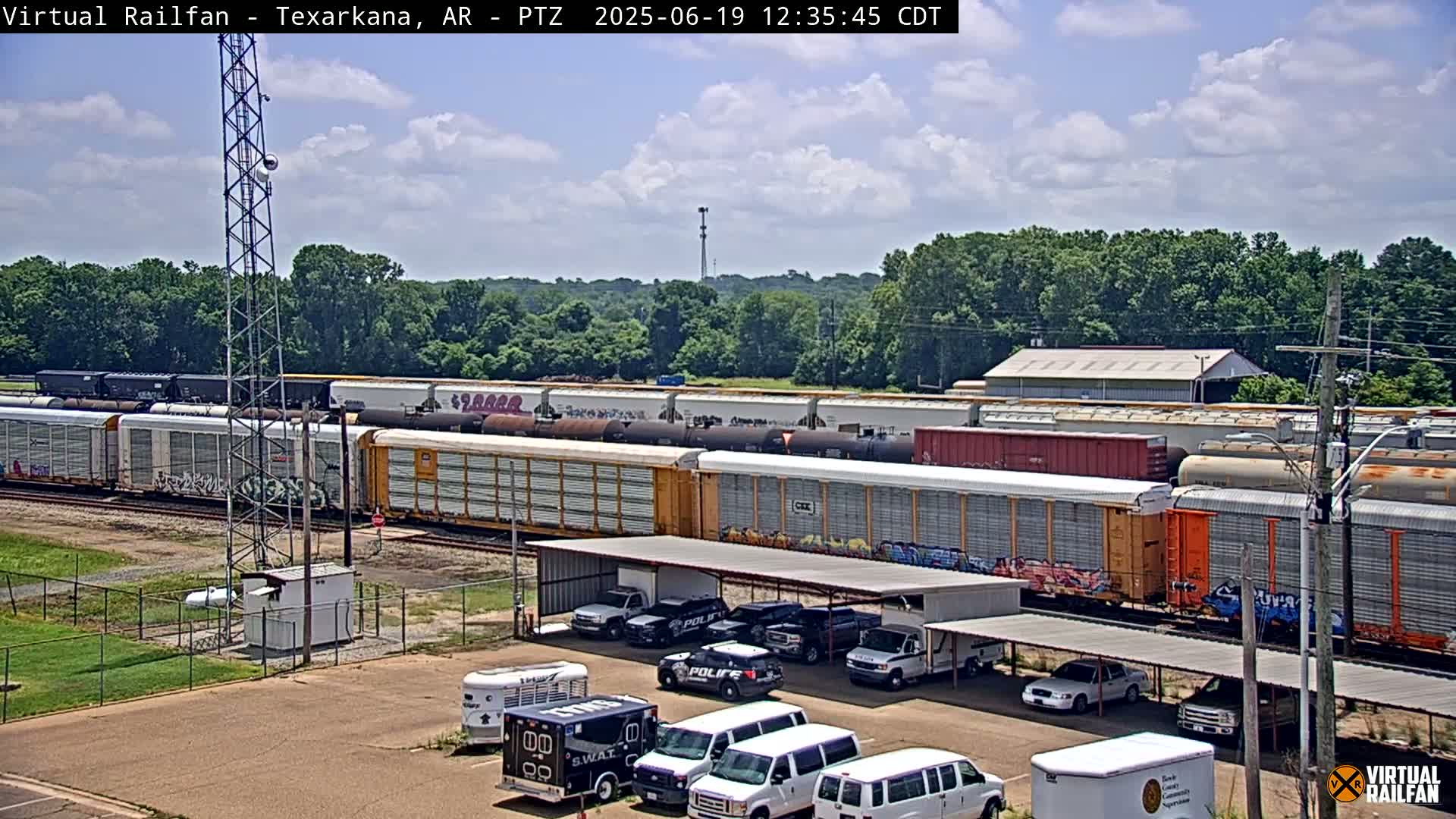 A long freight train sits on the tracks under a partly cloudy sky, with several police vehicles and vans parked in a lot nearby.