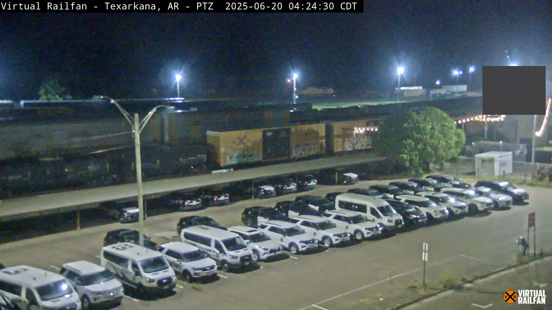 A long freight train is parked at night next to a large parking lot full of mostly white vehicles under bright lights.