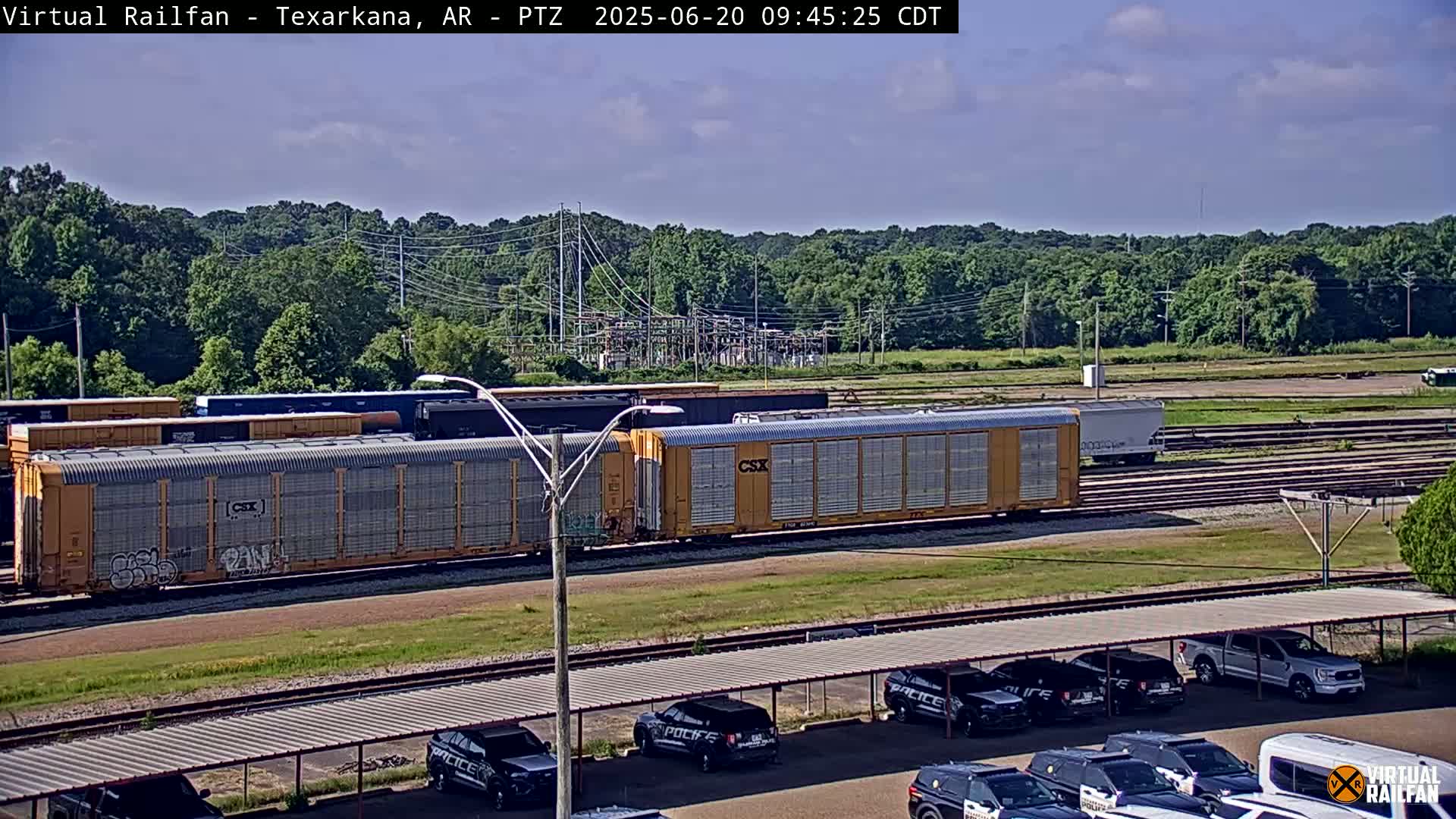 A mostly sunny day shows a train of yellow freight cars on a rail line next to a parking lot with several police cars under a covered structure, and a line of trees in the background.