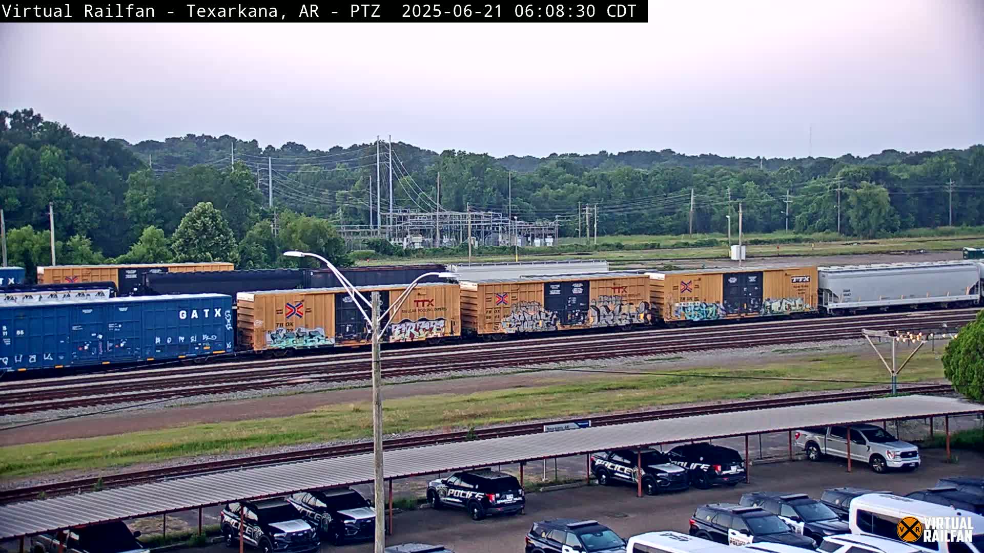 A freight train with graffiti-covered cars sits on the tracks next to a parking lot of police vehicles under a covered structure, all under a partly cloudy sky.