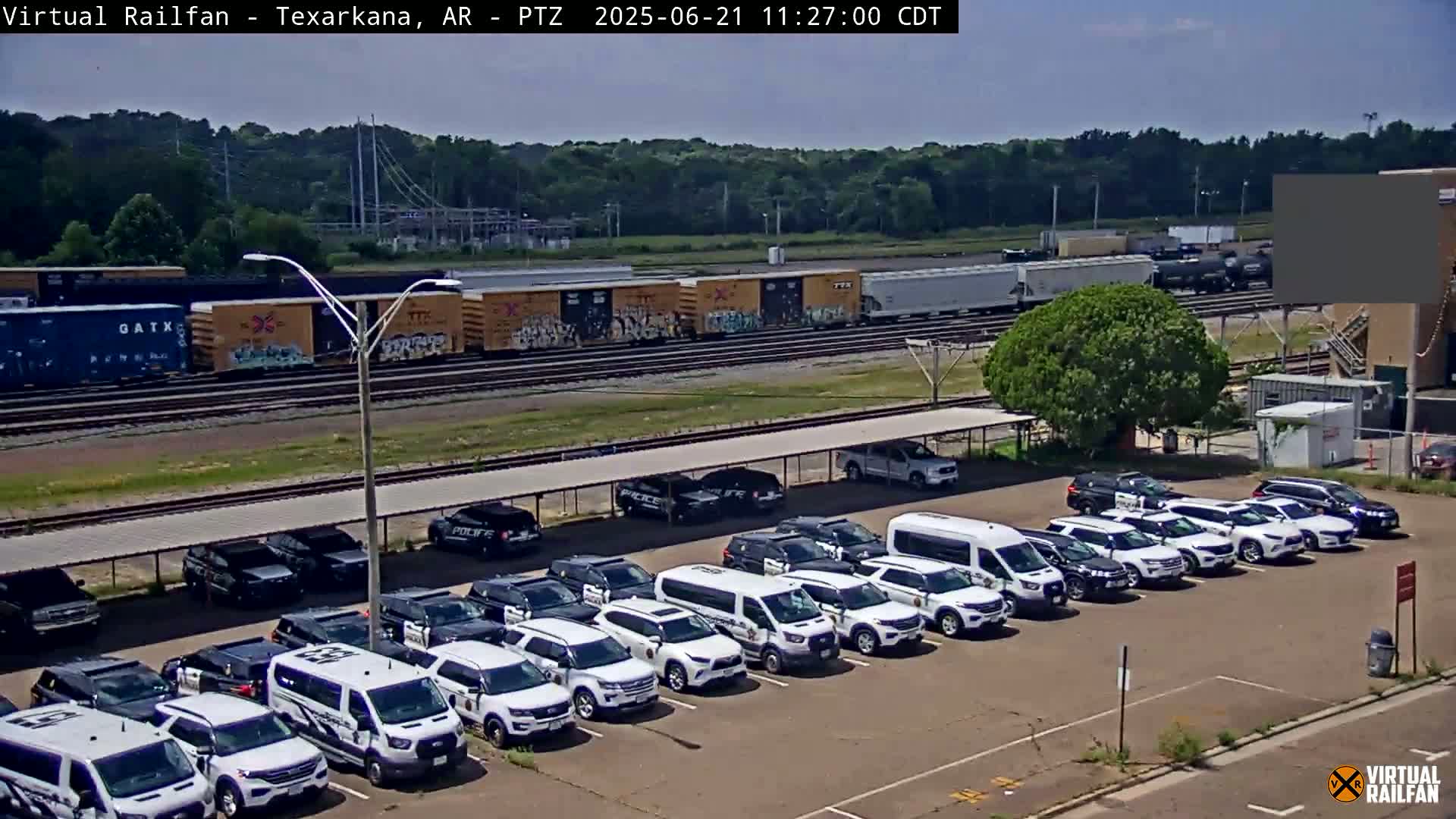 Under a sunny sky, a freight train passes by a parking lot full of police vehicles parked under a covered area.