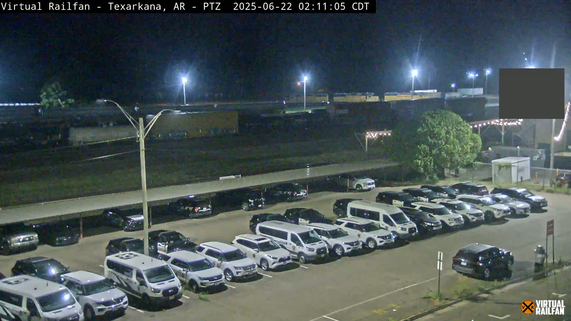 A nighttime view of a mostly empty parking lot filled with white vehicles under a covered structure, adjacent to a railway with a train in the distance.