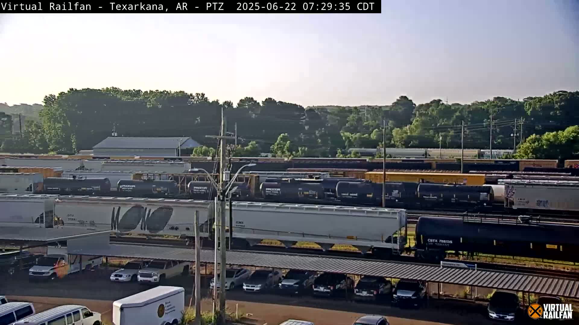 A long freight train passes through a rail yard on a partly sunny morning, with cars parked under a covered structure nearby.