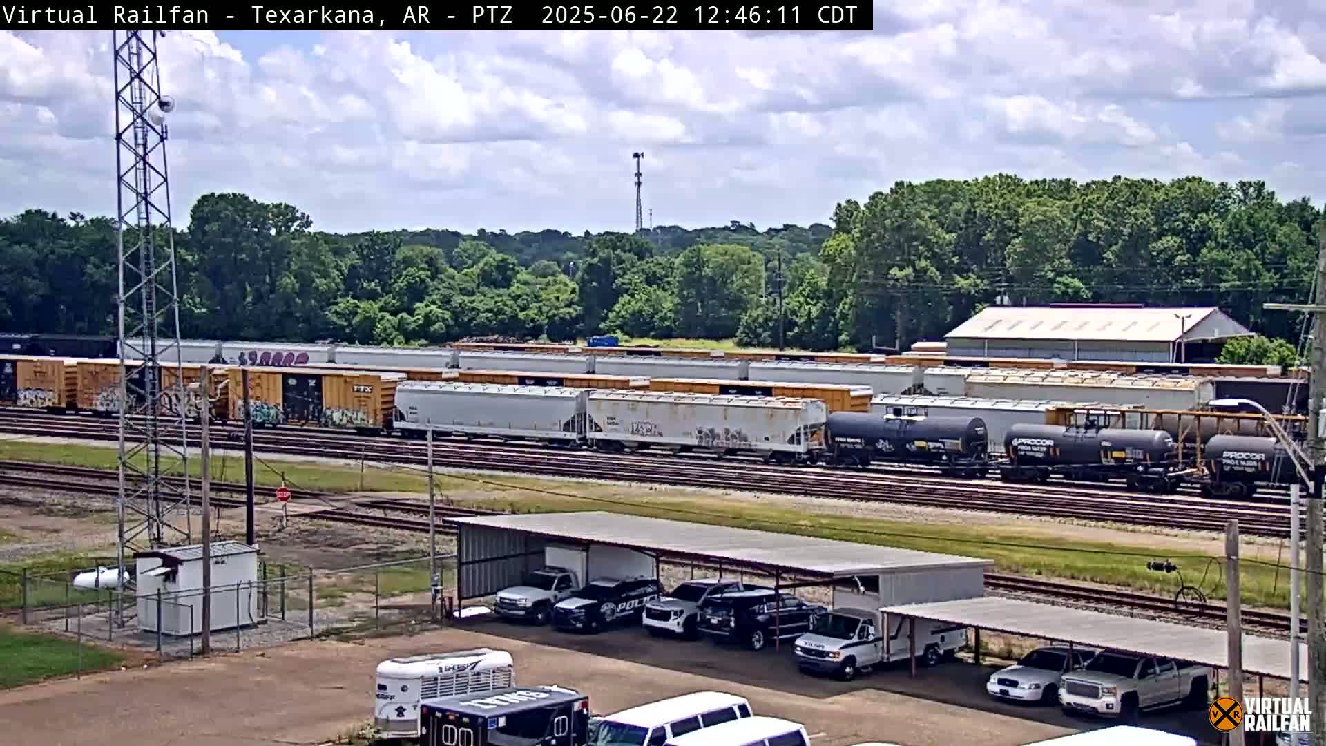 A long freight train sits on railroad tracks under a partly cloudy sky, with several police vehicles parked under a covered structure nearby.