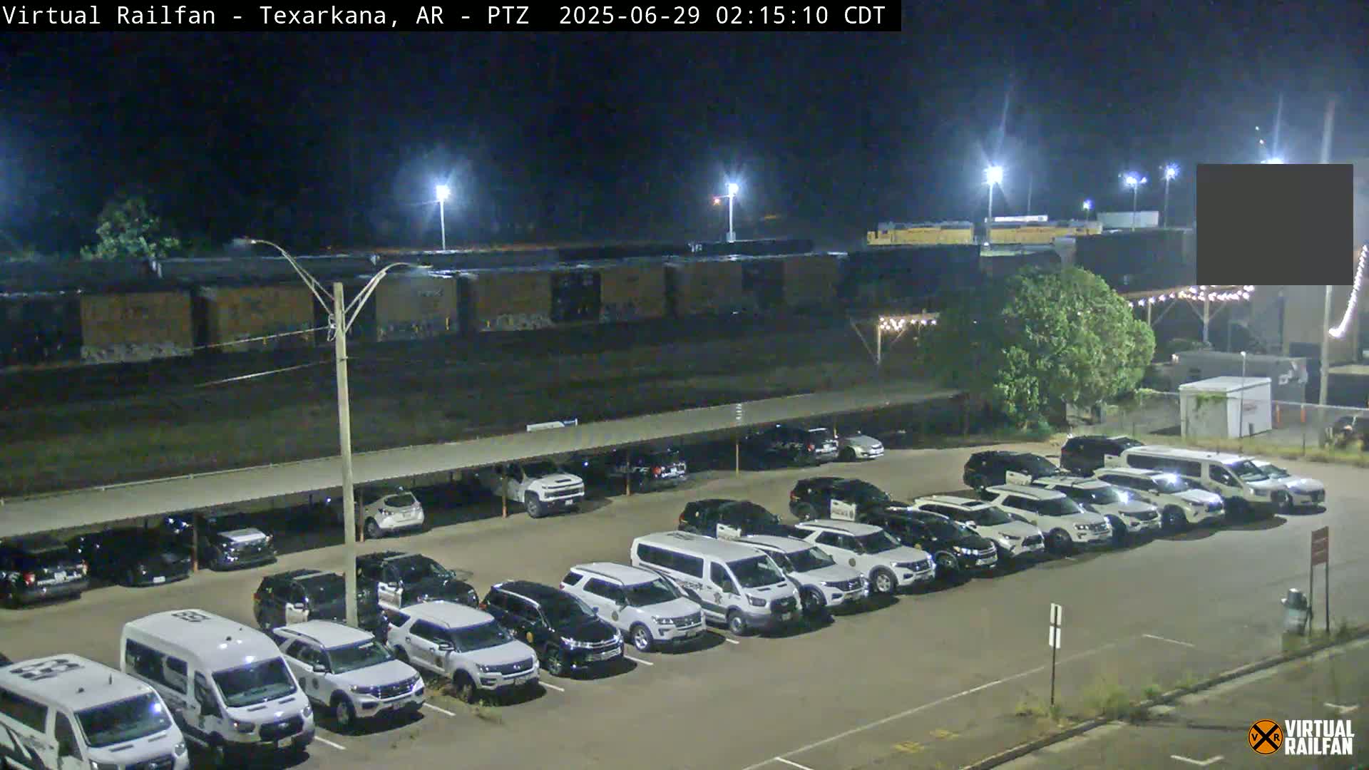 A nighttime view of a parking lot filled with numerous vehicles, mostly white SUVs and vans, under a covered structure next to a train yard.