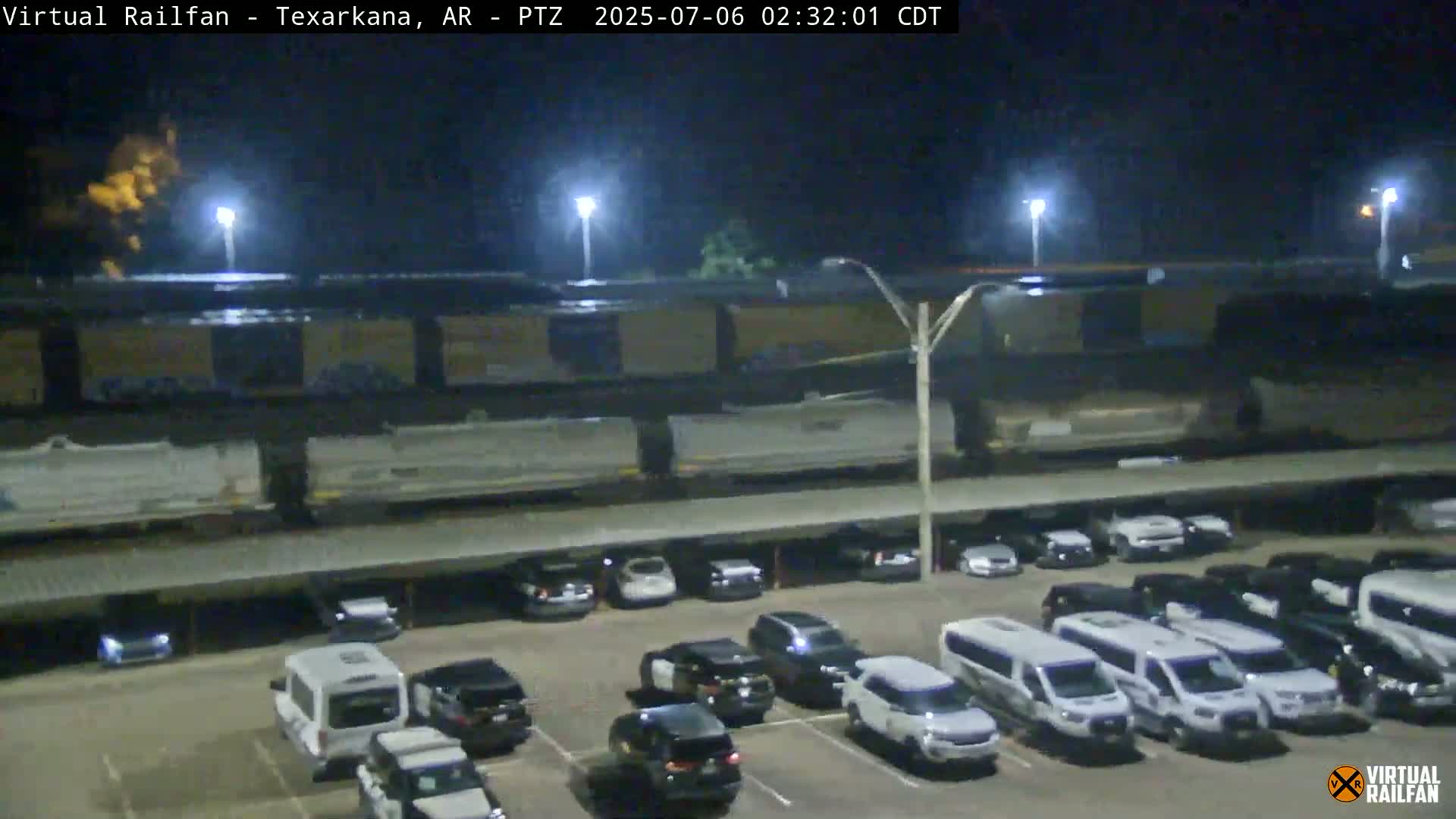 A nighttime view shows a long freight train parked near a parking lot full of cars and vans under a covered structure.