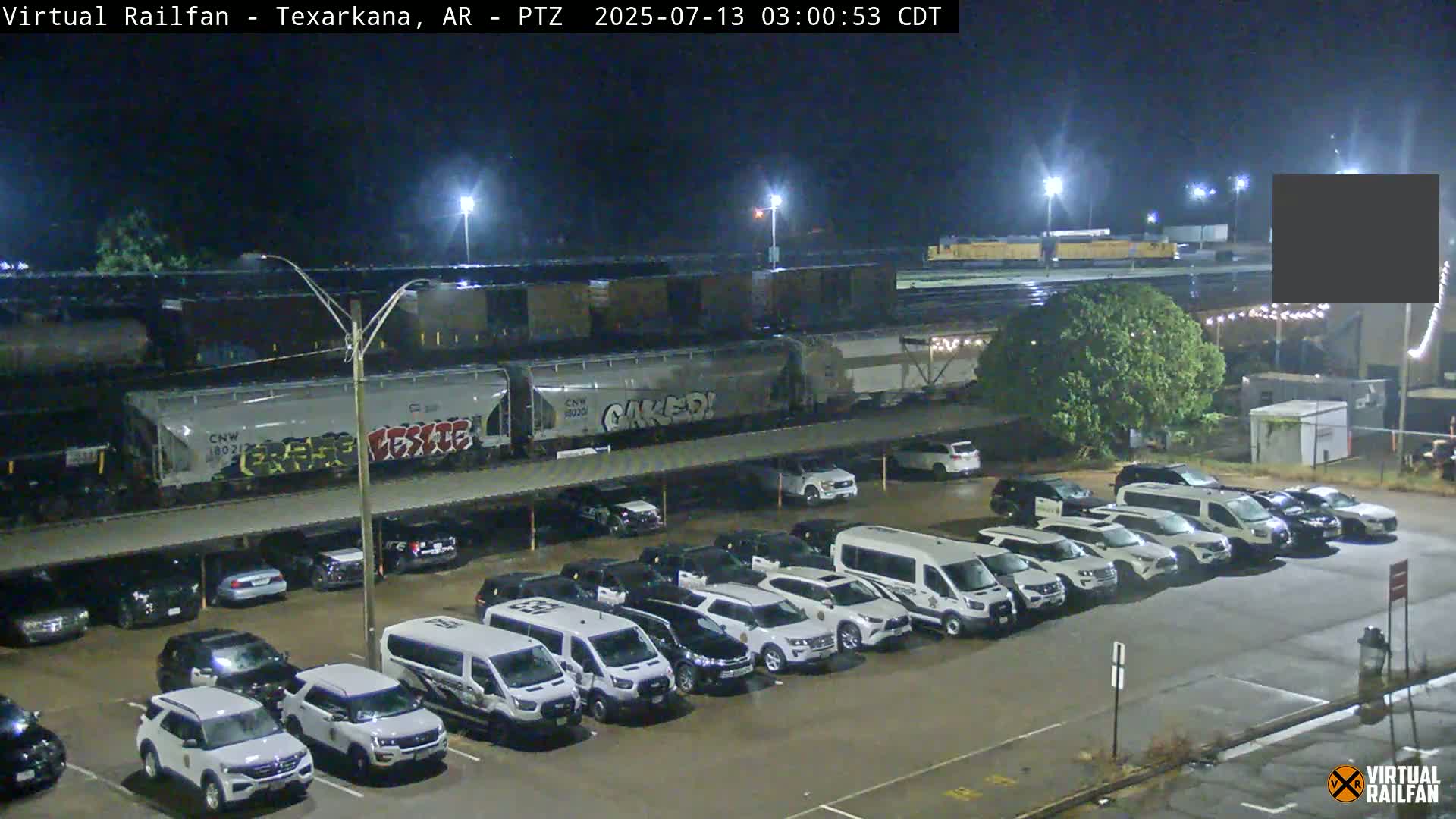 A freight train passes over a parking lot full of mostly white SUVs and vans at night under lightly overcast skies.