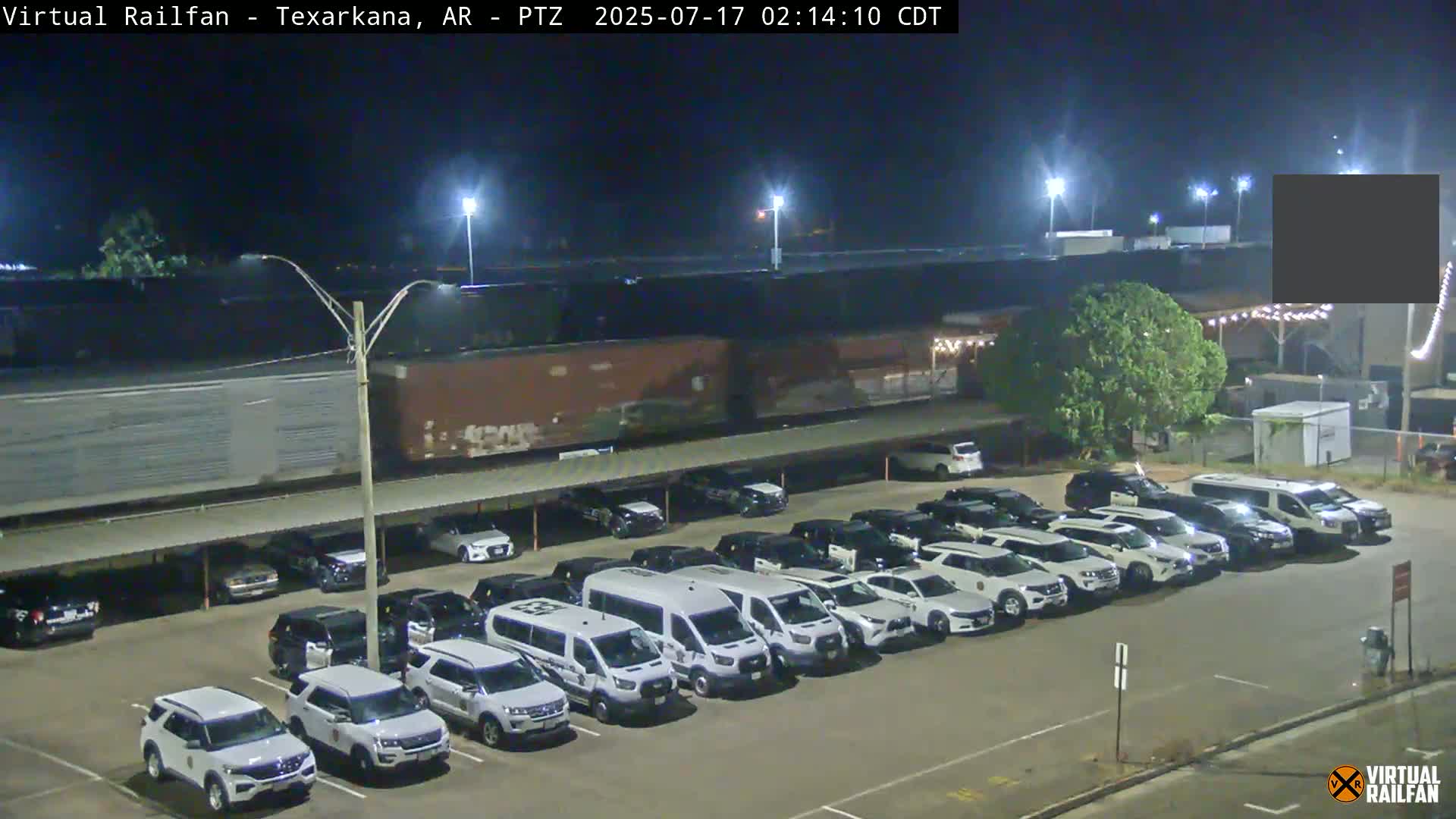 A long freight train passes behind a parking lot full of mostly white SUVs and vans at night under bright lights.