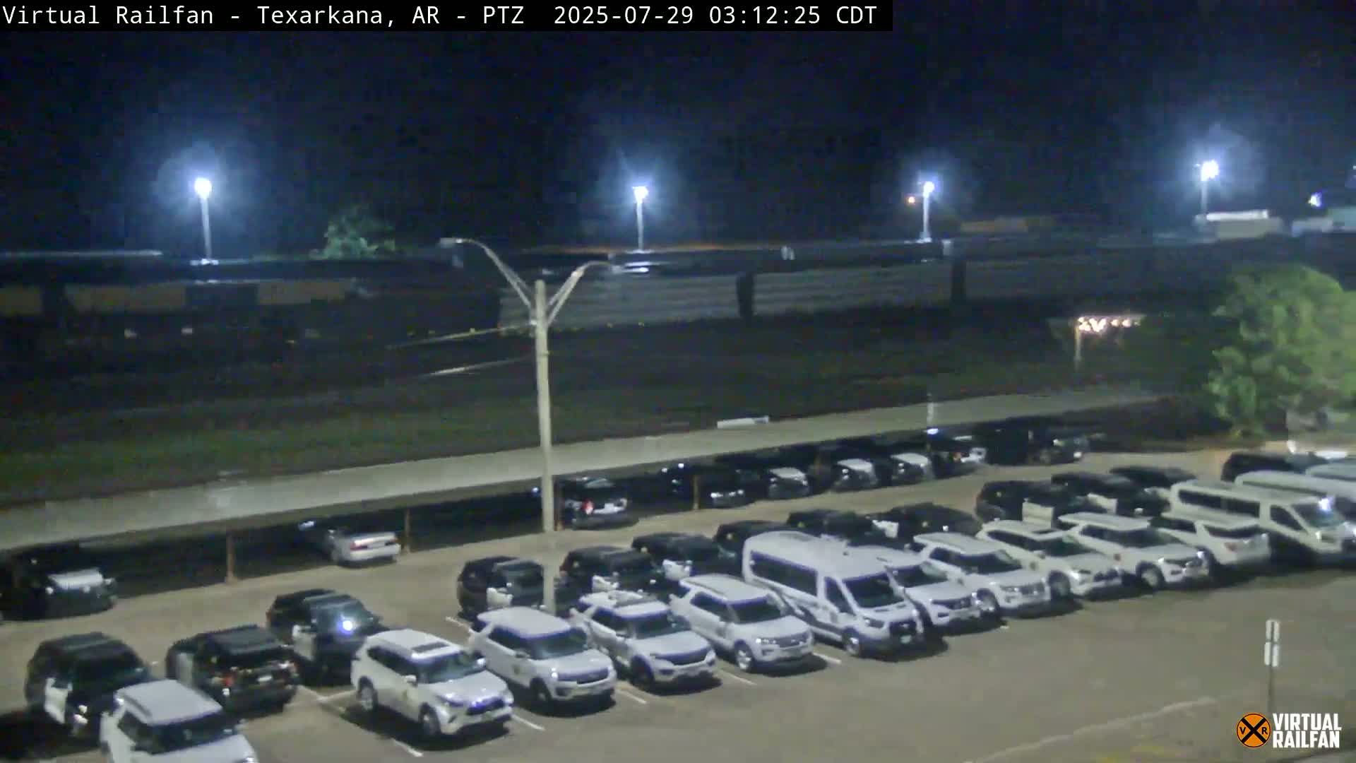 A long freight train passes by a parking lot full of mostly white vehicles at night under bright lights.