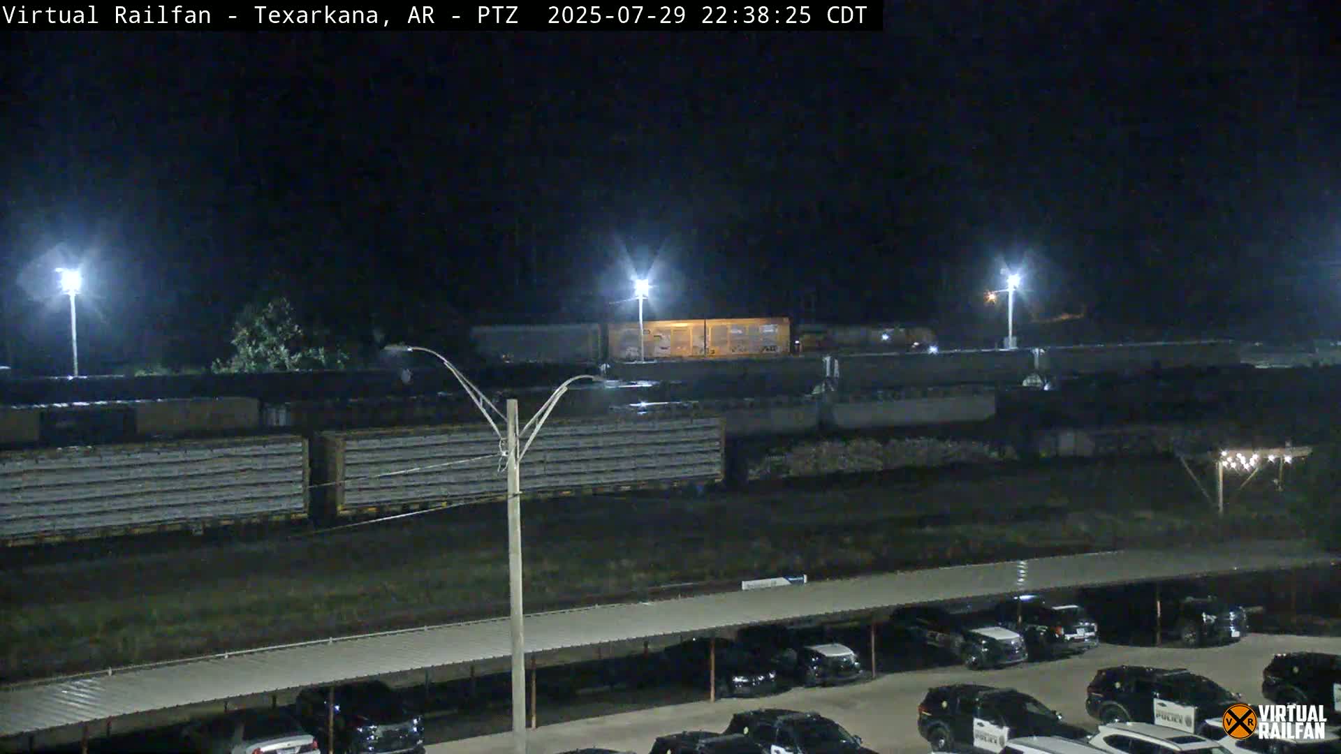 A long freight train passes by a parking lot full of mostly white vehicles at night under bright lights.