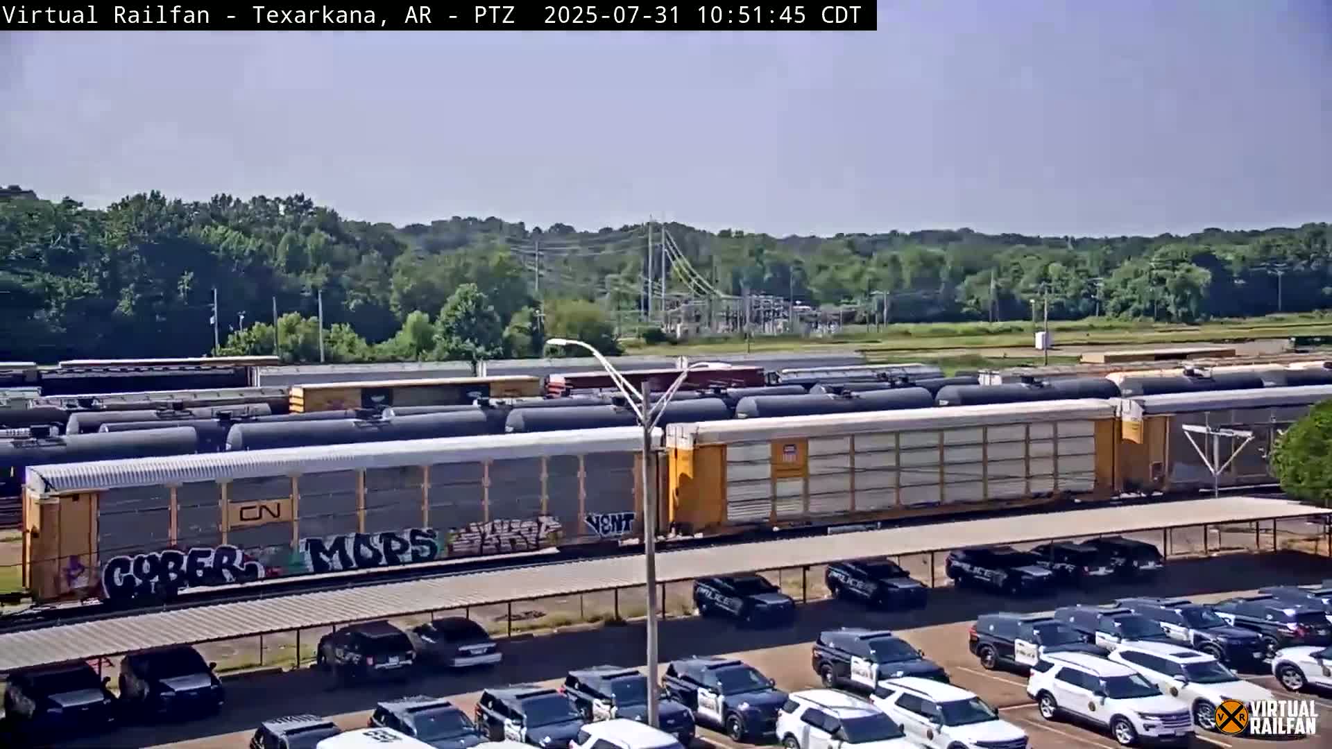 A long freight train with graffiti sits in a rail yard next to a parking lot full of police vehicles under a clear, sunny sky.