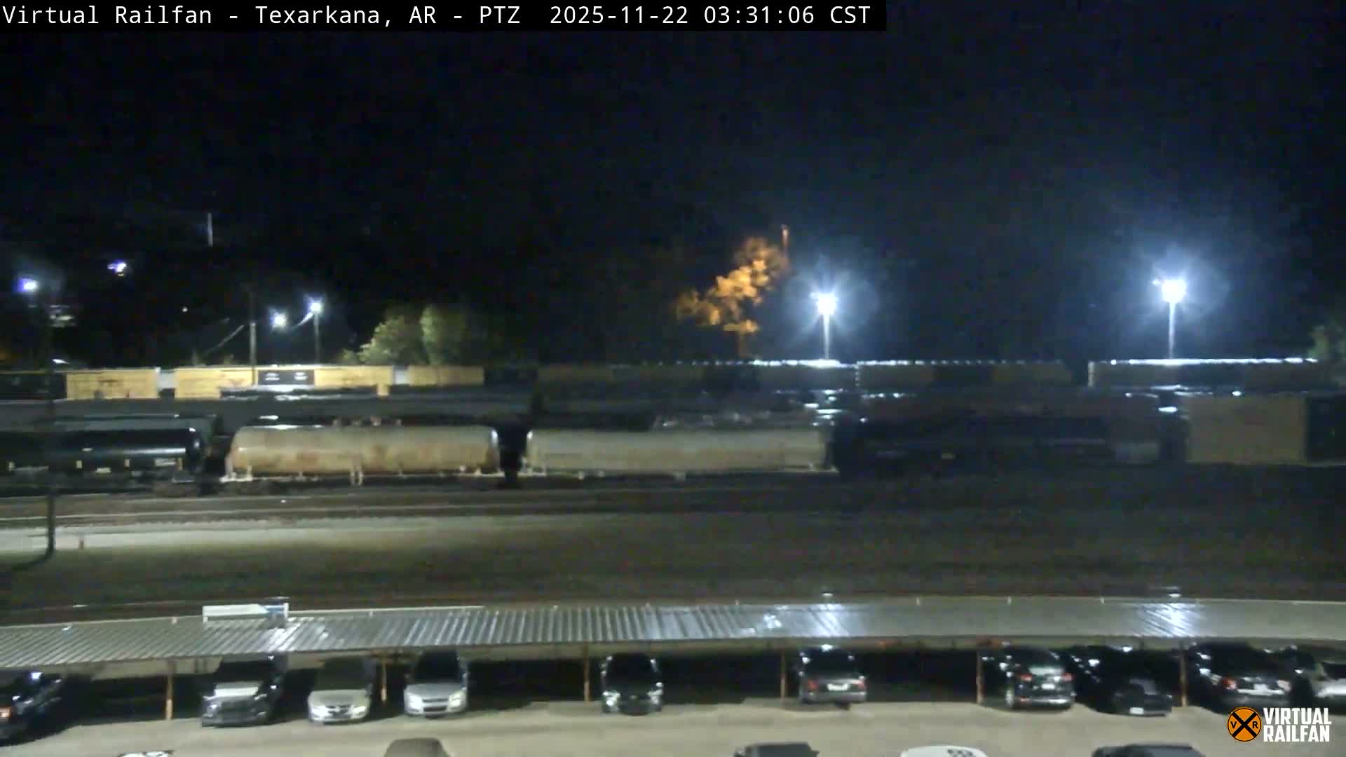 On a clear night, a wide view captures a train yard filled with various railcars under the glow of numerous artificial lights, with a parking lot and several parked cars in the foreground.