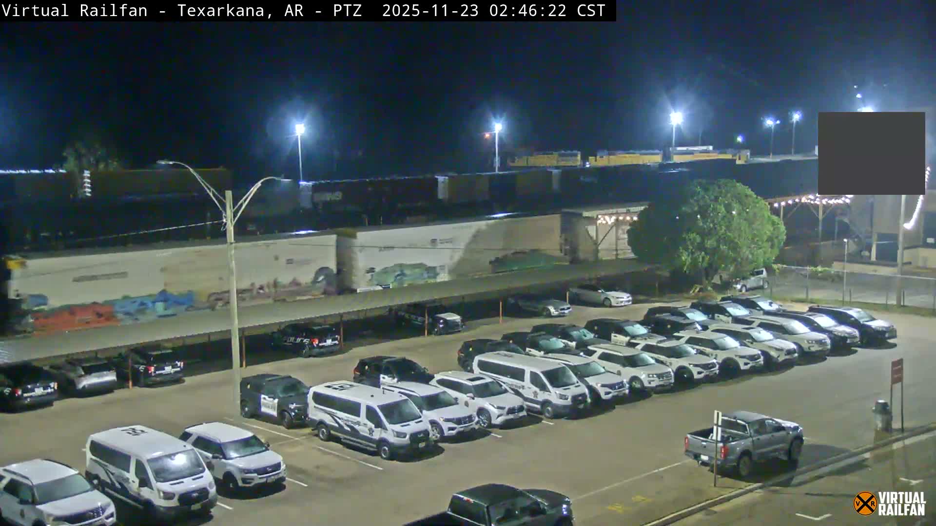 A clear night reveals a brightly lit outdoor parking lot densely packed with various vehicles, including several white fleet vans and SUVs, situated below an elevated railway line where freight cars are visible, with distant industrial buildings.