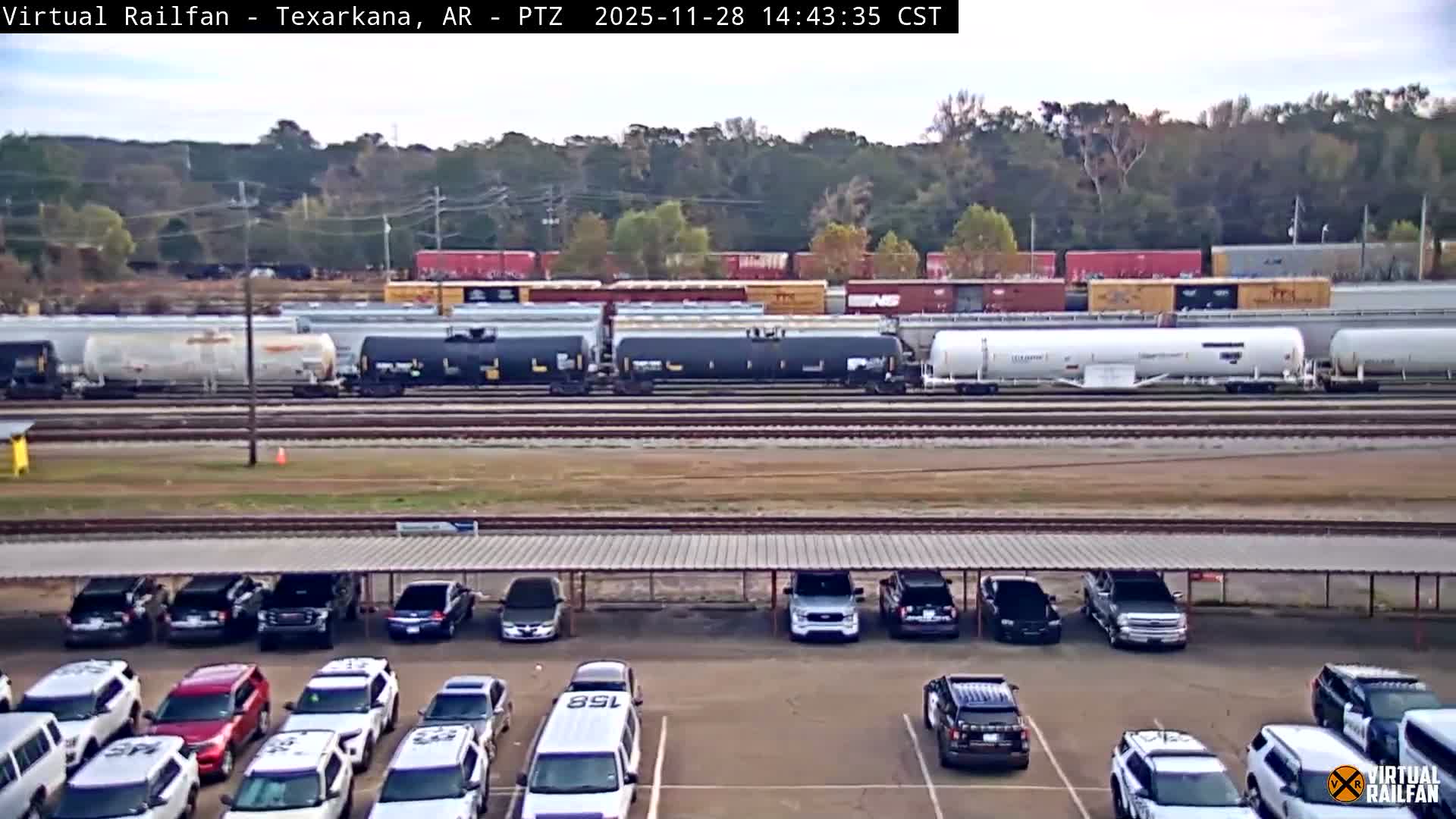 A wide outdoor view shows numerous freight trains with various tank cars and boxcars on multiple tracks, with a parking lot full of cars in the foreground, all under a bright, partly cloudy sky.