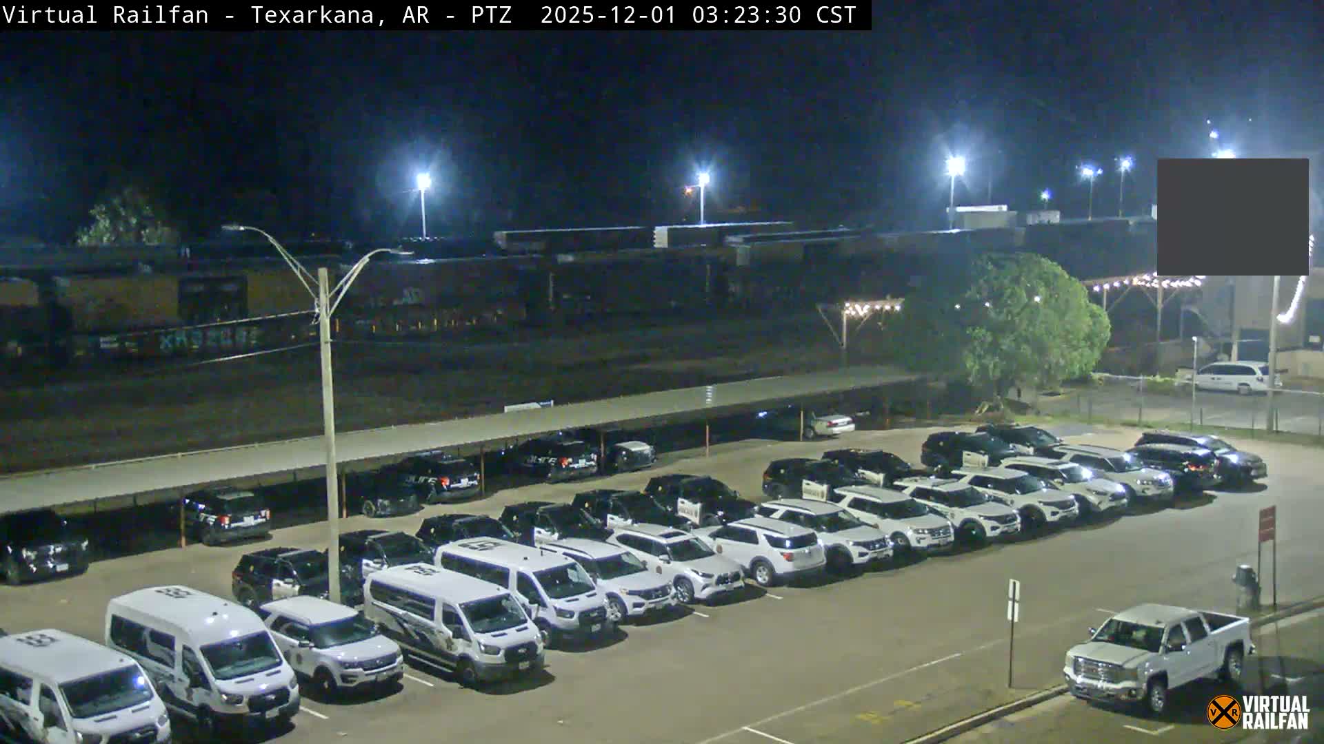 The image depicts a brightly lit outdoor parking lot at night with clear skies, filled with a mix of white vans and black and white SUVs (many resembling police vehicles), bordered by active train tracks with visible freight cars in the distance.