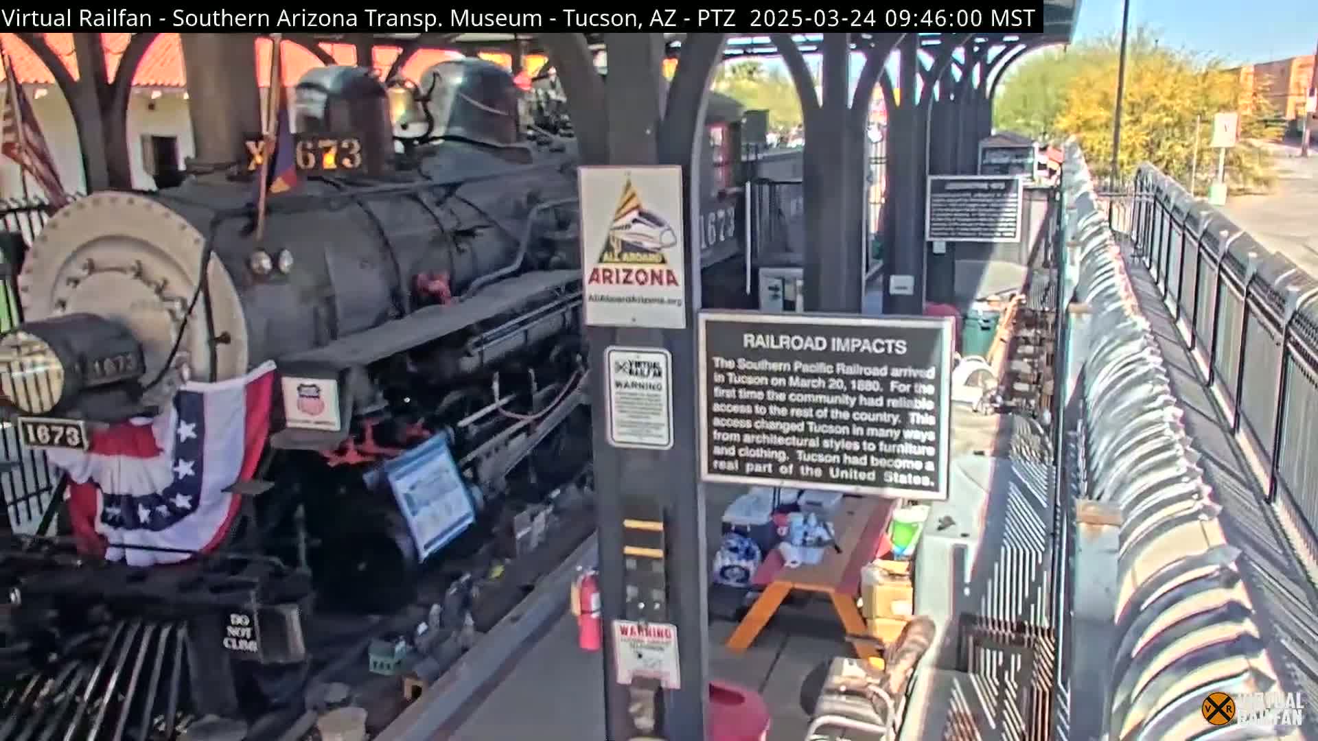 A dark-colored steam locomotive with American and Arizona flags adorns it sits under a covered structure on a sunny day.
