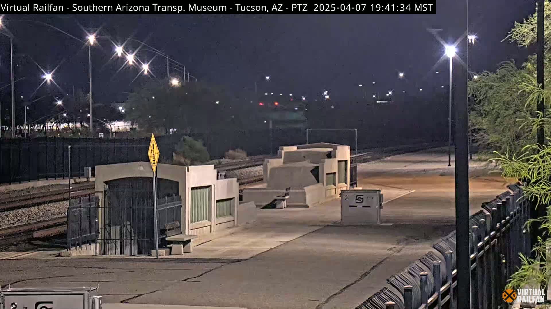 A nighttime view of a deserted train platform with concrete structures, a fence, and several lights illuminating the area.